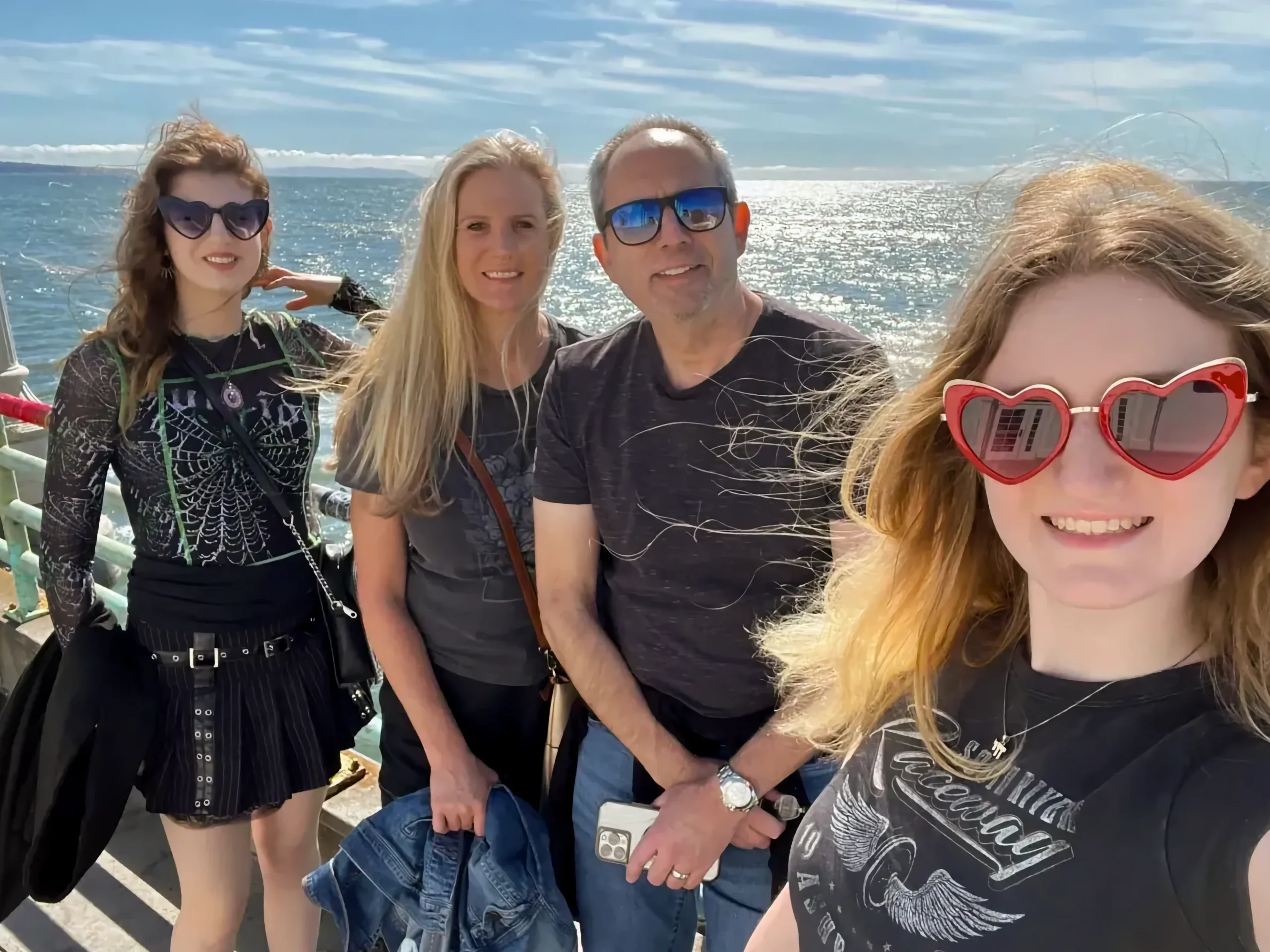 Allyson and Aharon Kessary takes a sunny selfie on a pier overlooking the bright blue water with their two daughters. They are wearing sunglasses, and the wind is blowing their hair.