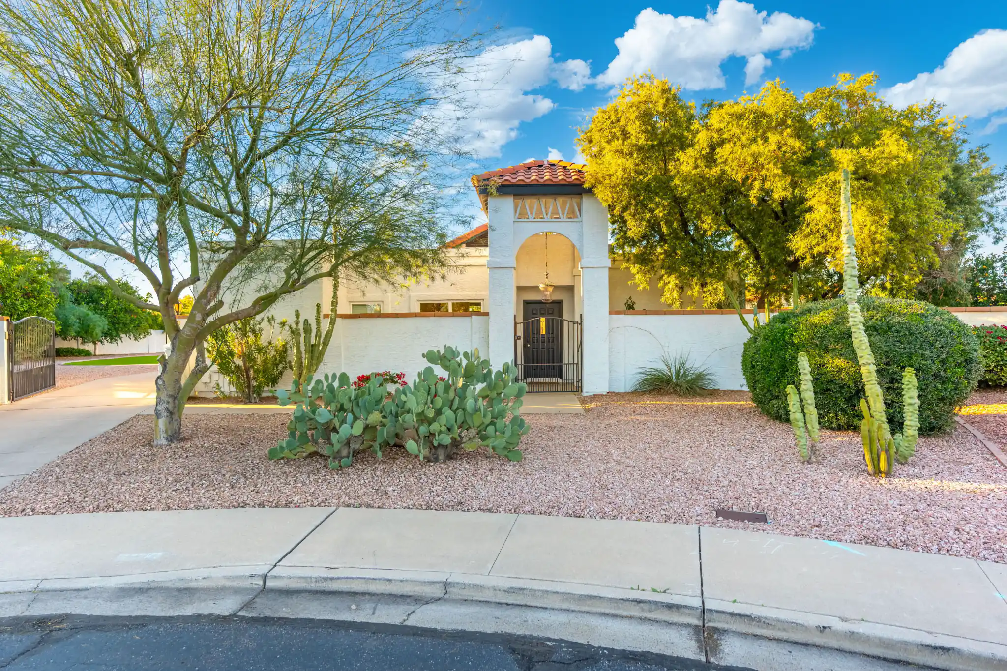 A white stucco home features an arched entryway with a tiled roof. The front yard is gravel, landscaped with a prickly pear cactus and desert trees.