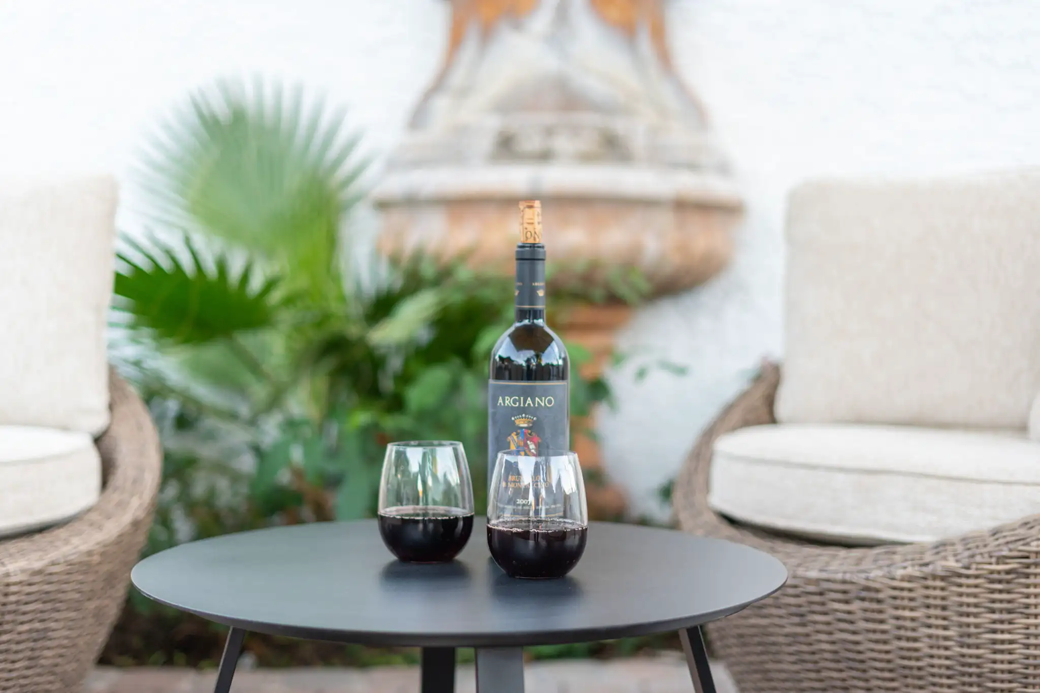 An outdoor patio setting features a bottle of red wine and two glasses full of wine resting on a dark table between two wicker chairs with cream cushions.
