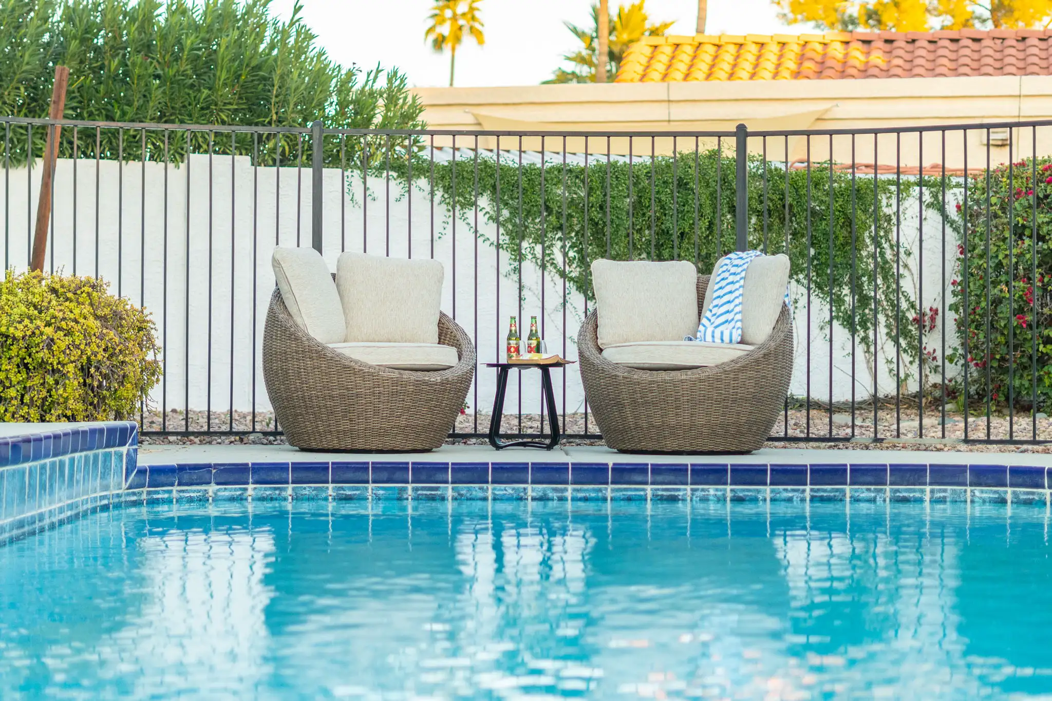 Two curved wicker chairs with light cushions face a blue swimming pool. A side table holds two beers, suggesting relaxation. The chairs sit on the pool deck near a black fence.