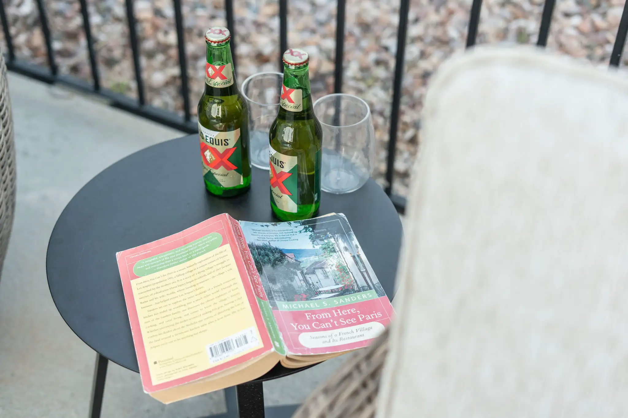 An open book and two bottles of Dos Equis beer with glasses sit on a small black patio table, implying a relaxing afternoon reading.