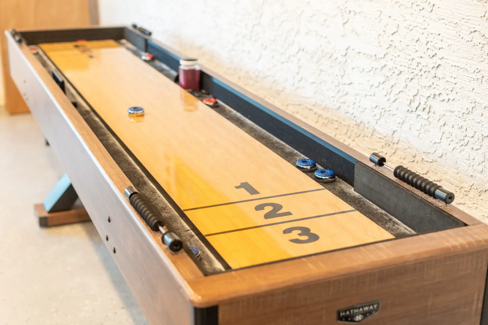 A close-up view of a wooden shuffleboard table with scoring lanes marked 1, 2, and 3. Several blue and red pucks are on the playing surface.