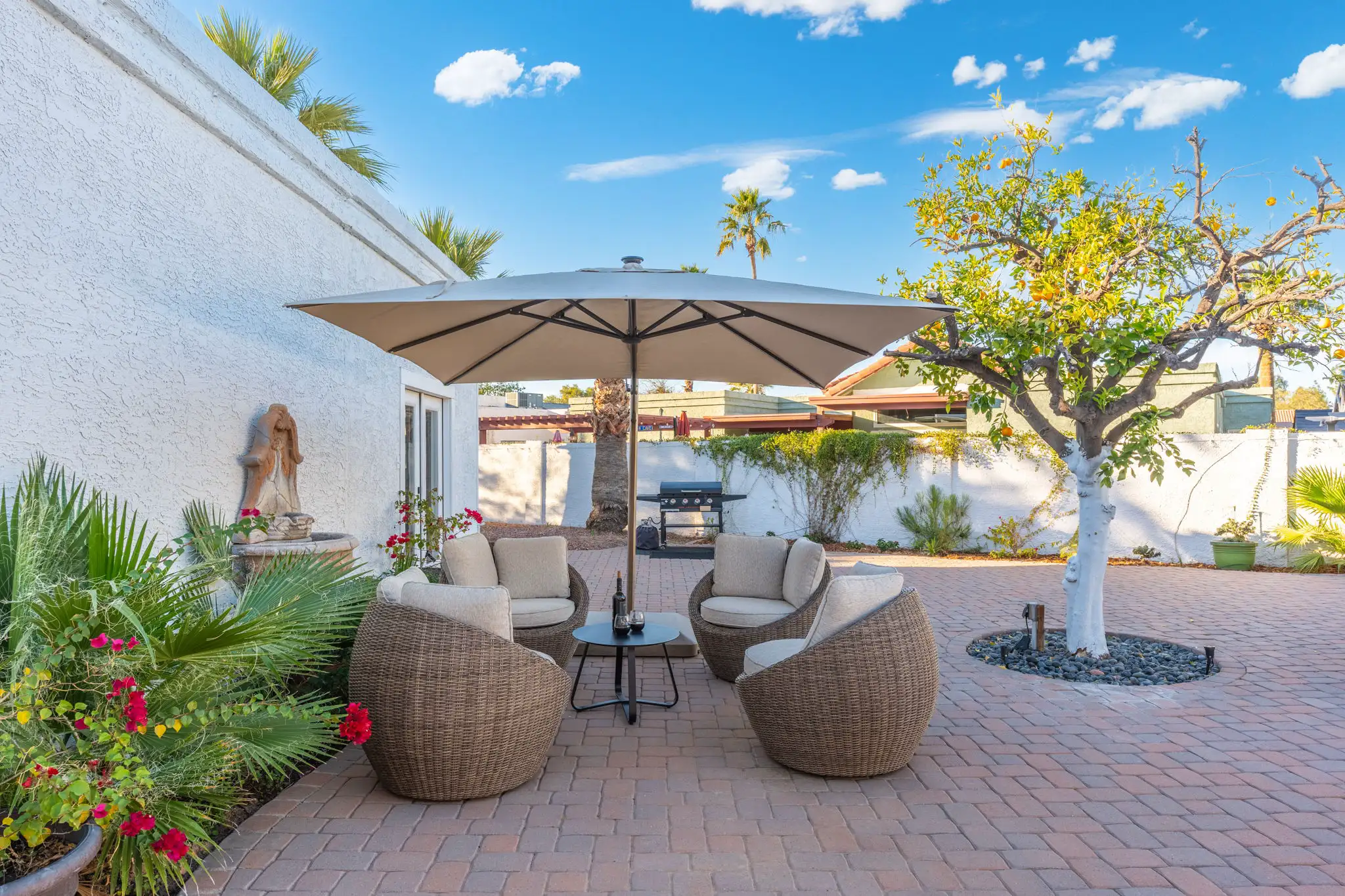 Four wicker patio chairs and a table sit under a large umbrella on a brick paver patio, surrounded by lush plants, a stone fountain, and a productive citrus tree.