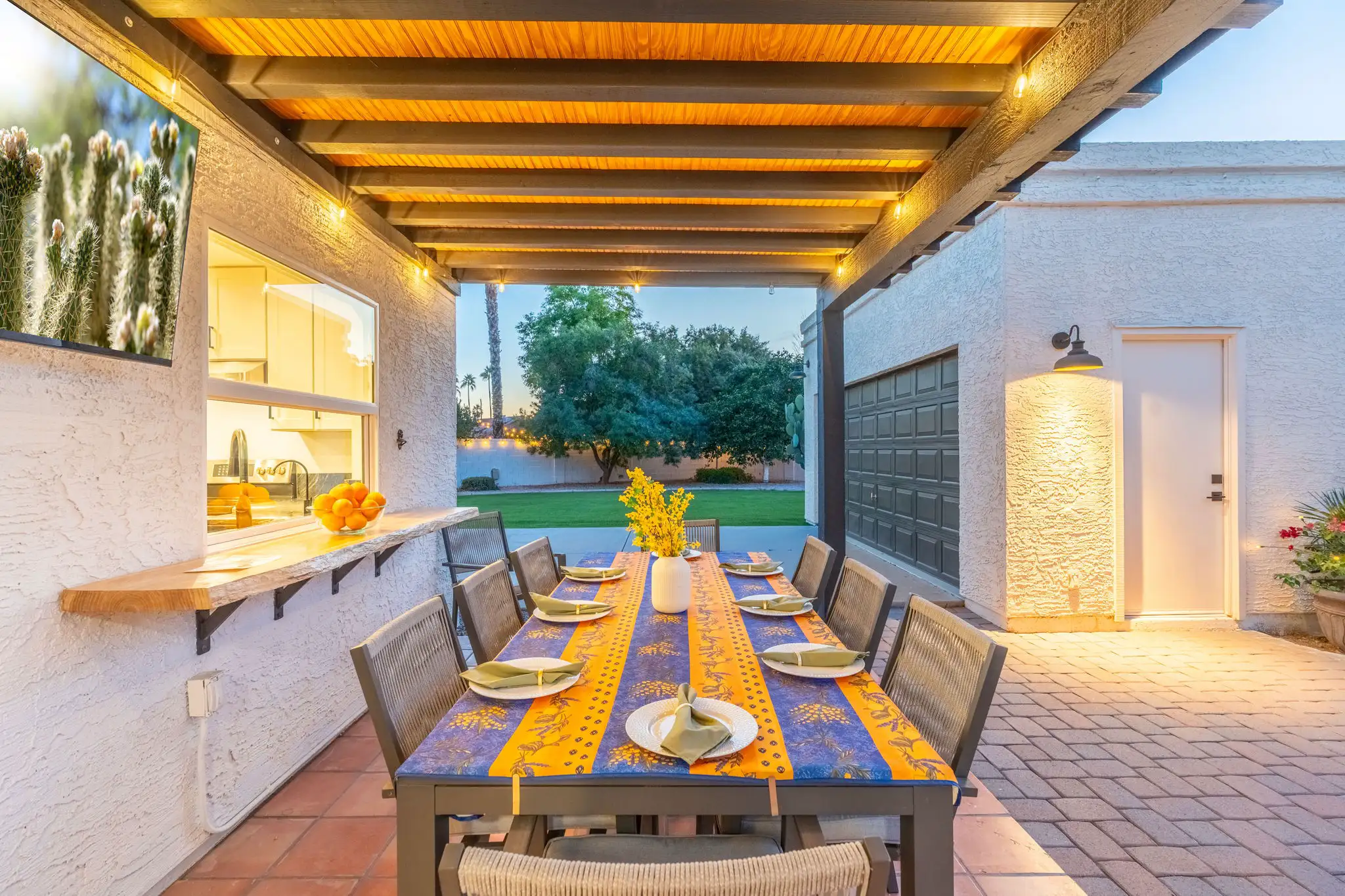 An inviting covered outdoor dining area features a table set for eight with a colorful runner. String lights and a kitchen pass-through illuminate the space at dusk.