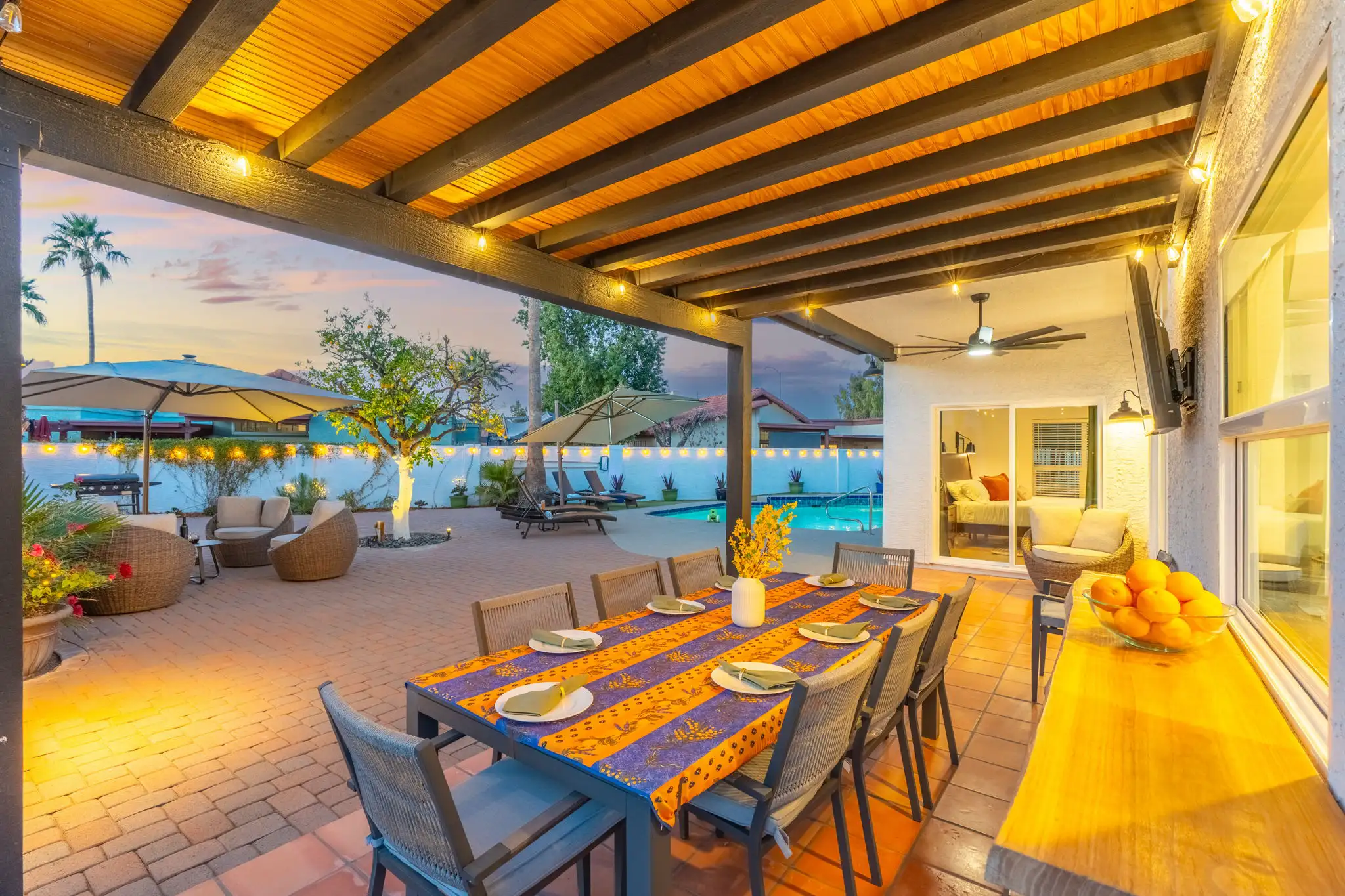 An inviting covered patio at twilight. A long, set dining table sits under a wooden ceiling and string lights, next to a comfortable seating area and a pool.