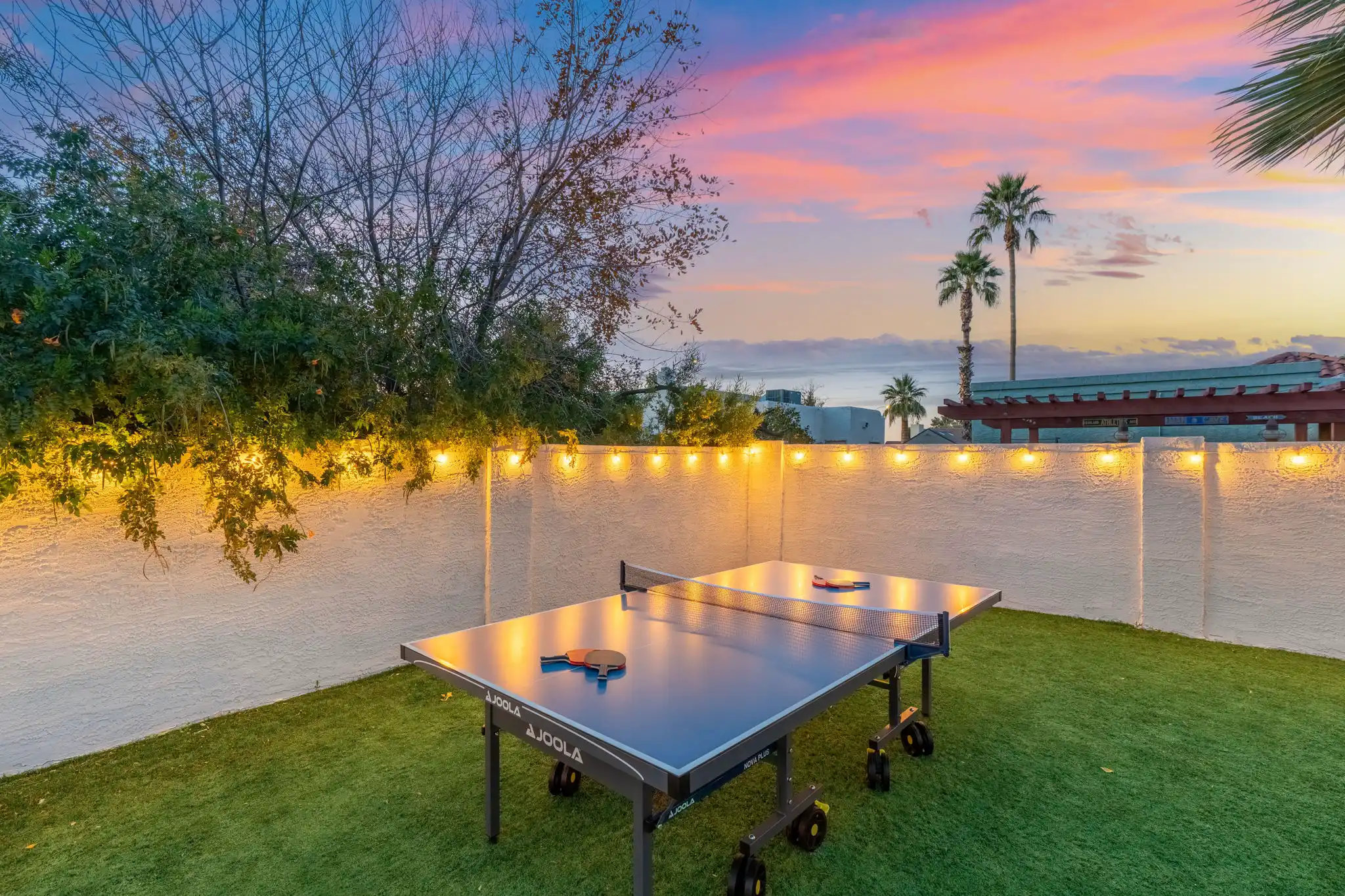 An outdoor table tennis setup rests on green artificial turf in a yard, illuminated by string lights on a white stucco wall. The colorful sunset sky features palm trees in the background.