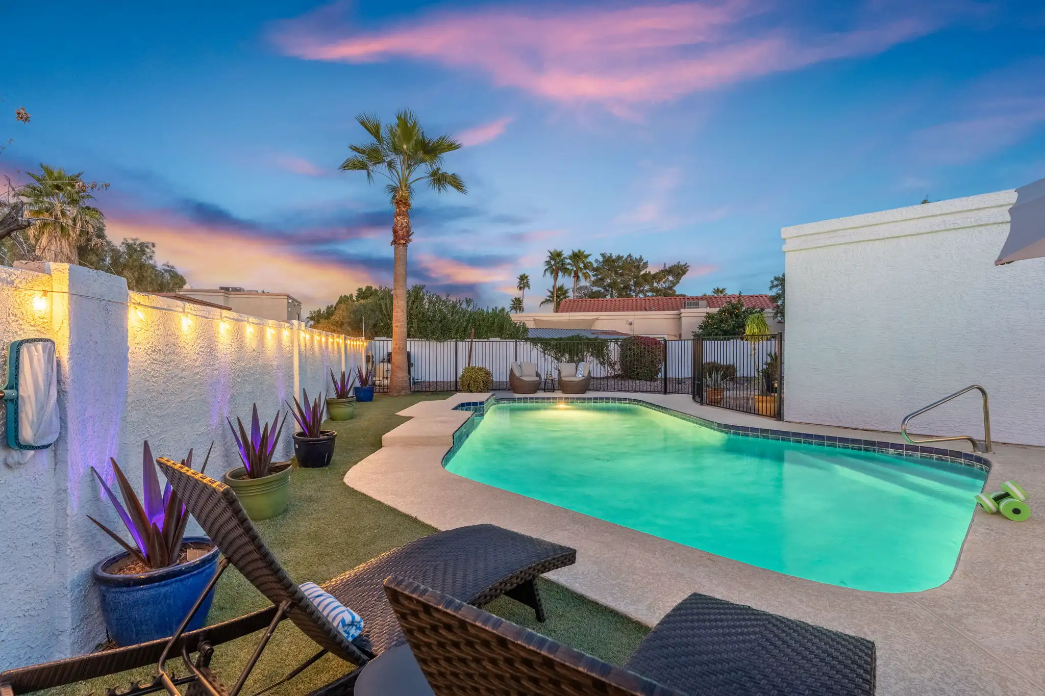 A resort-style swimming pool at twilight, surrounded by turf, a tall palm tree, and wicker lounge chairs. String lights illuminate the white stucco wall.