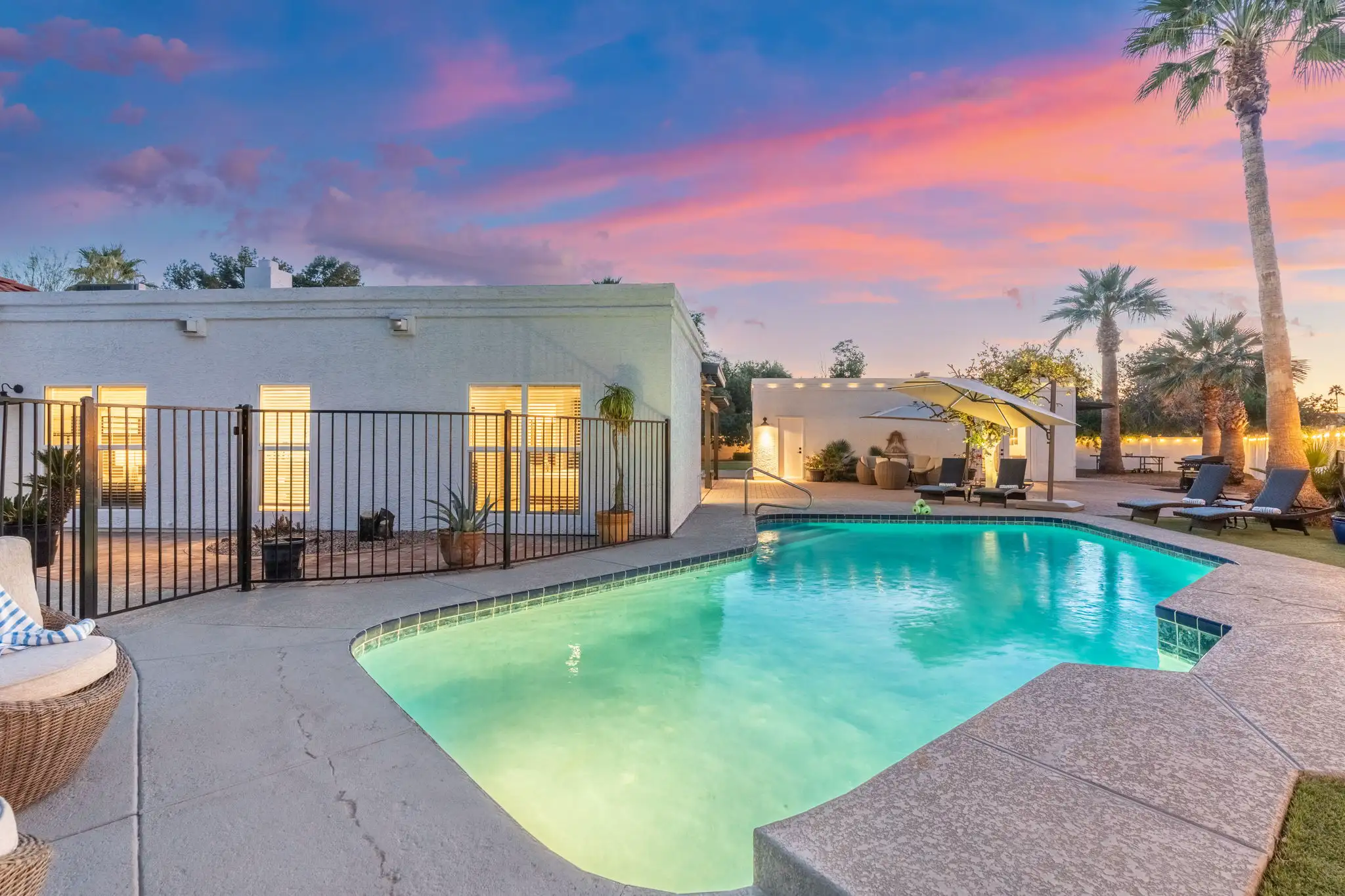 A vacation rental backyard featuring a brightly lit swimming pool, lounge chairs, and palm trees under a stunning pink and blue twilight sky.