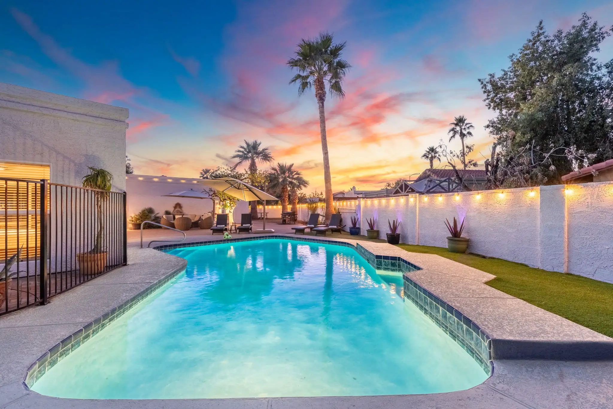 A bright blue swimming pool surrounded by a patio with lounge chairs and a tall palm tree. The backyard is framed by string-lit white walls under a vibrant sunset sky.