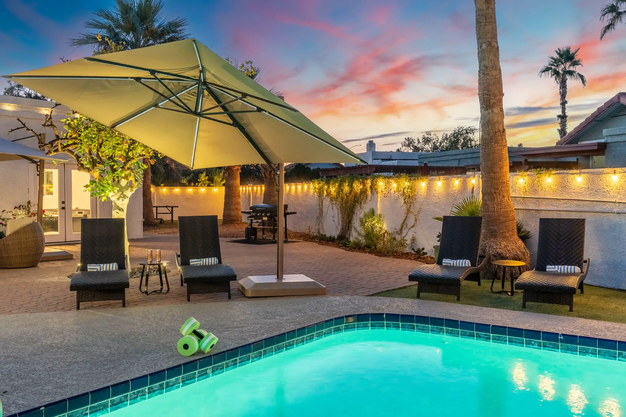 A tropical pool deck at dusk with a large illuminated umbrella casting light over four lounge chairs. The vibrant turquoise water contrasts with the colorful sunset sky.