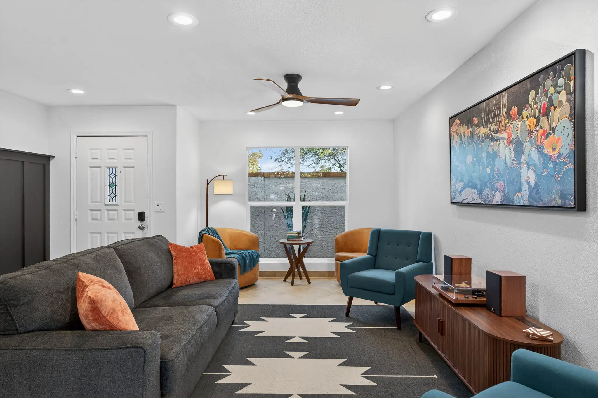 A bright, modern living room with recessed lighting, a dark gray sofa, a geometric rug, and a colorful cactus art print above a curved wood console.
