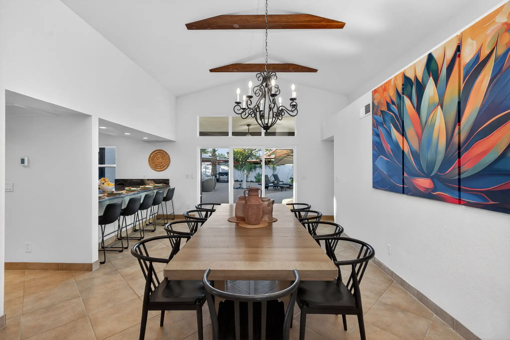 A bright dining room with a vaulted ceiling, wood beams, and a black chandelier. A long wooden table is surrounded by black chairs, adjacent to a bar and a patio entrance.