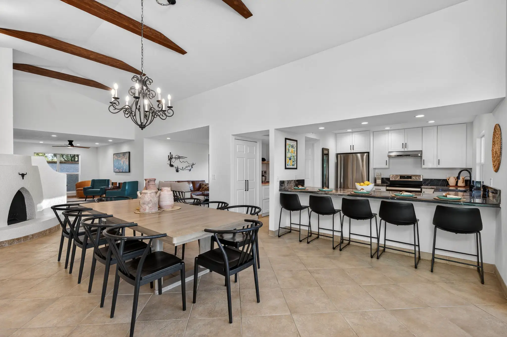 A bright open concept room featuring a wooden dining table and a white kitchen bar with black stools. The space has beige tile floors and a vaulted ceiling with dark wood beams.