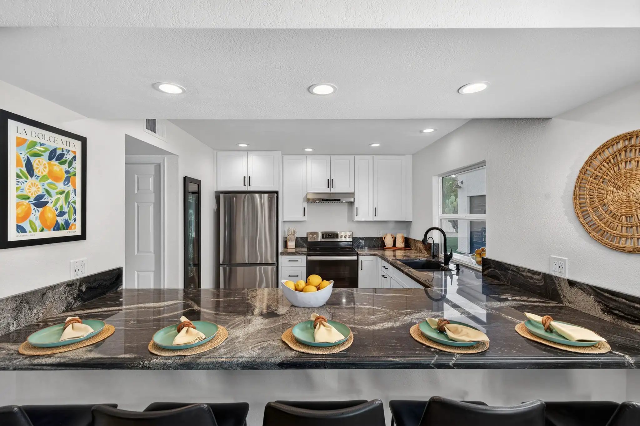 A clean, modern white kitchen with stainless appliances is viewed over a dark granite bar top set with five plates and a bowl of lemons.