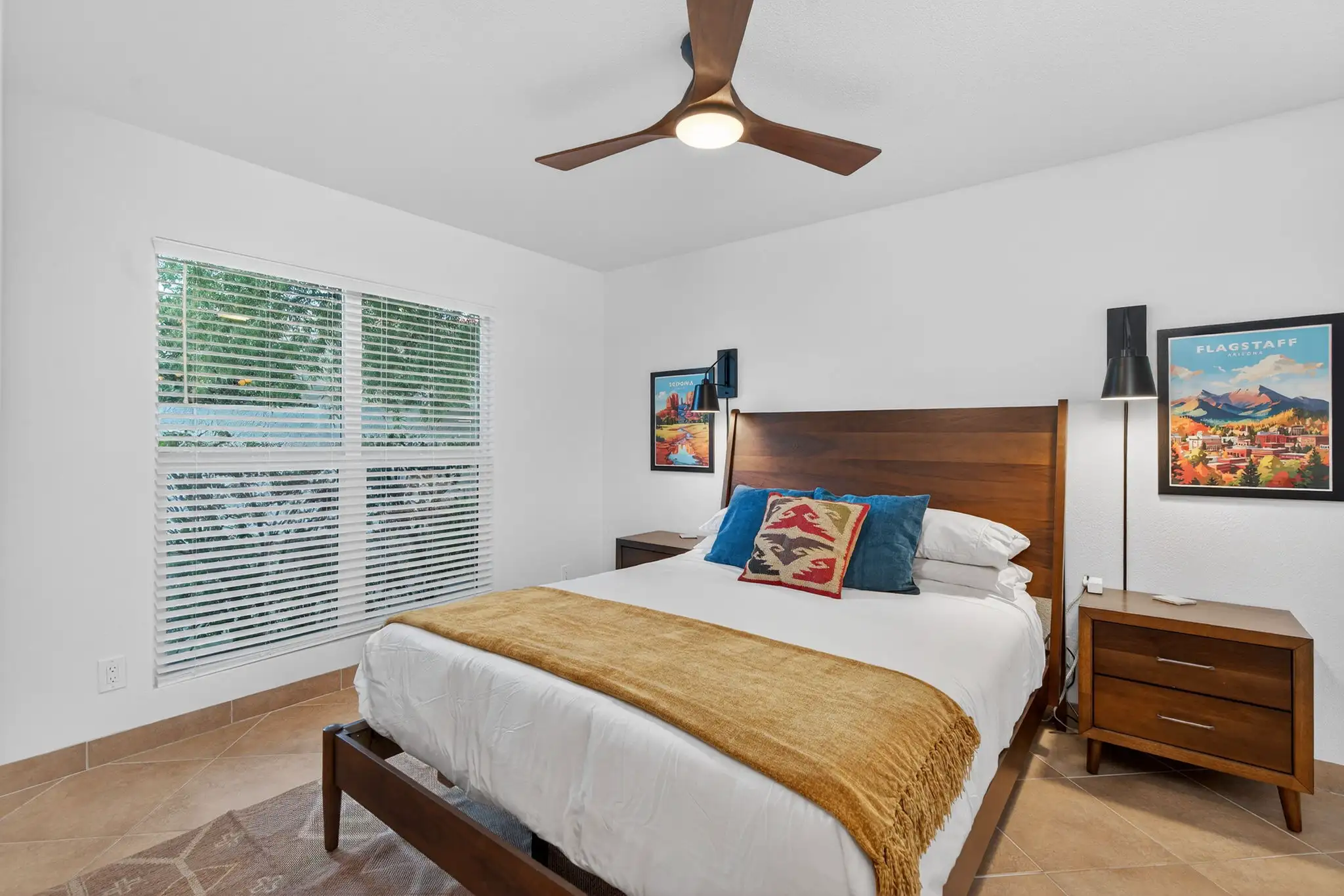 A spacious, modern bedroom featuring a bed with white linens and a yellow throw blanket, flanked by nightstands and Arizona travel posters.