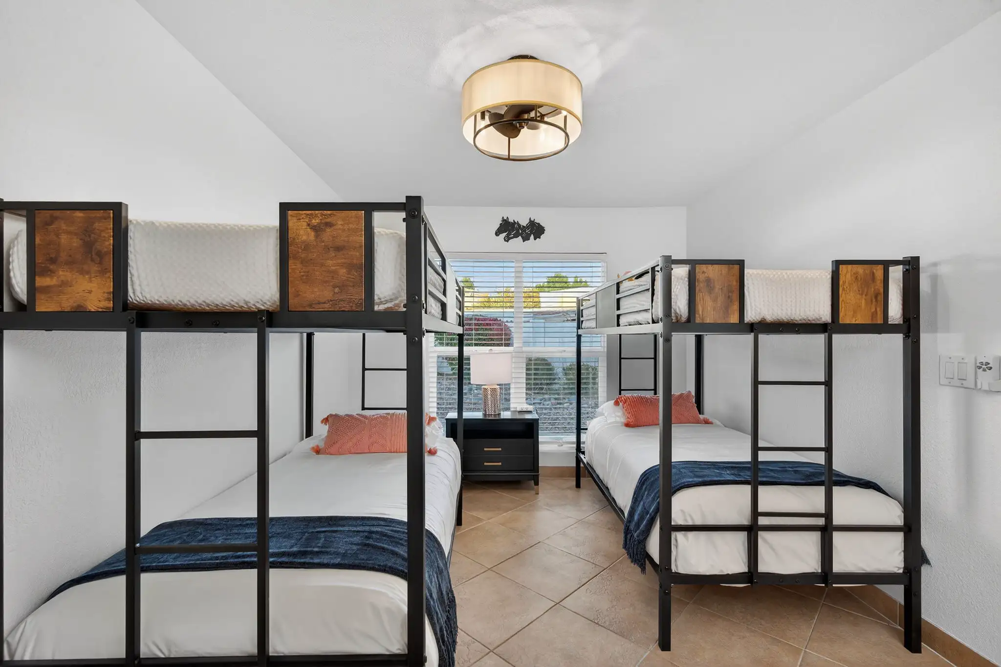A clean, bright bedroom with tile flooring featuring two black metal and wood bunk beds dressed with white and blue linens. A nightstand and lamp sit between the beds under a window.