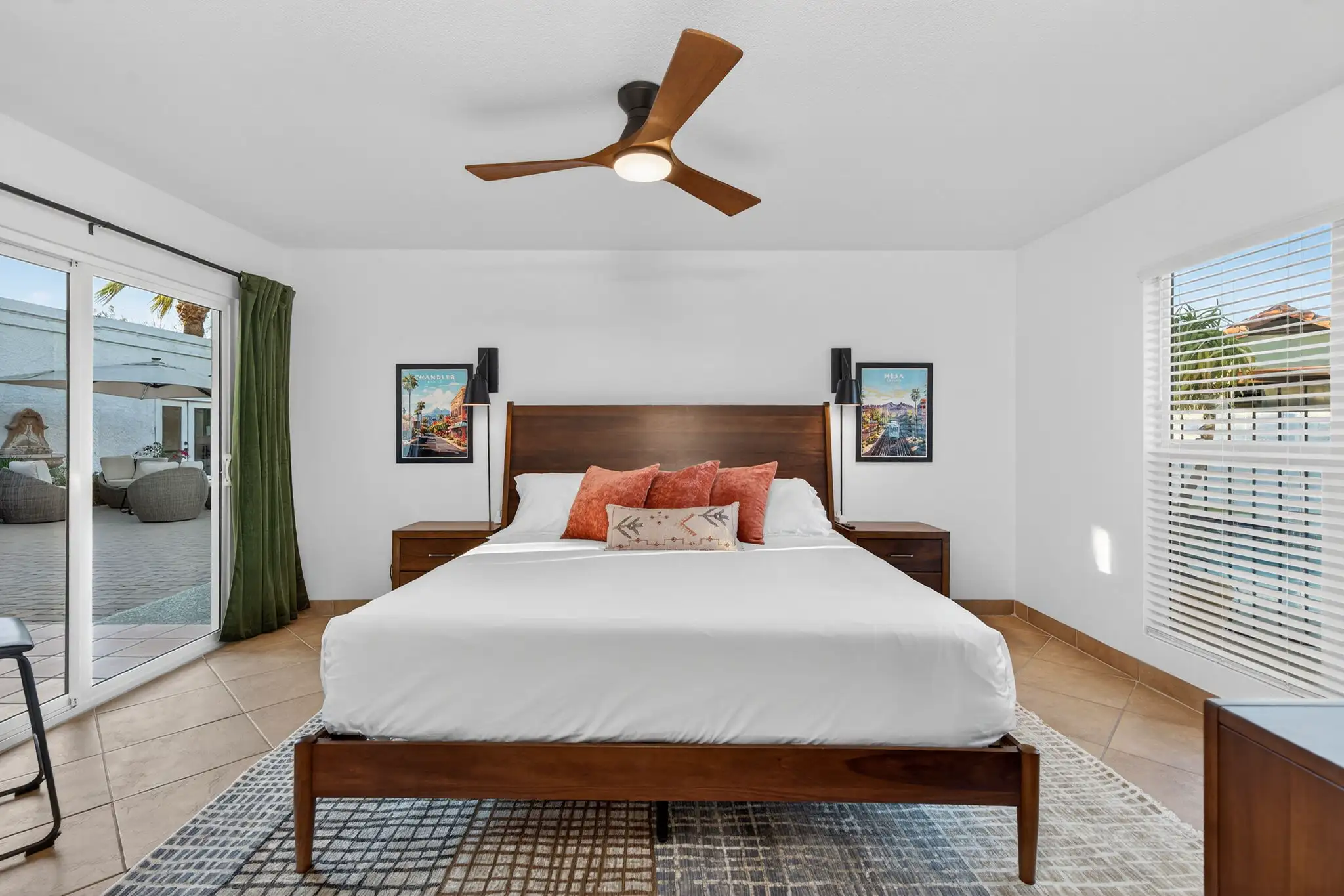A modern bedroom features a wooden bed with white linens and warm-toned pillows, a ceiling fan, and sliding glass doors that open to a sunlit patio.