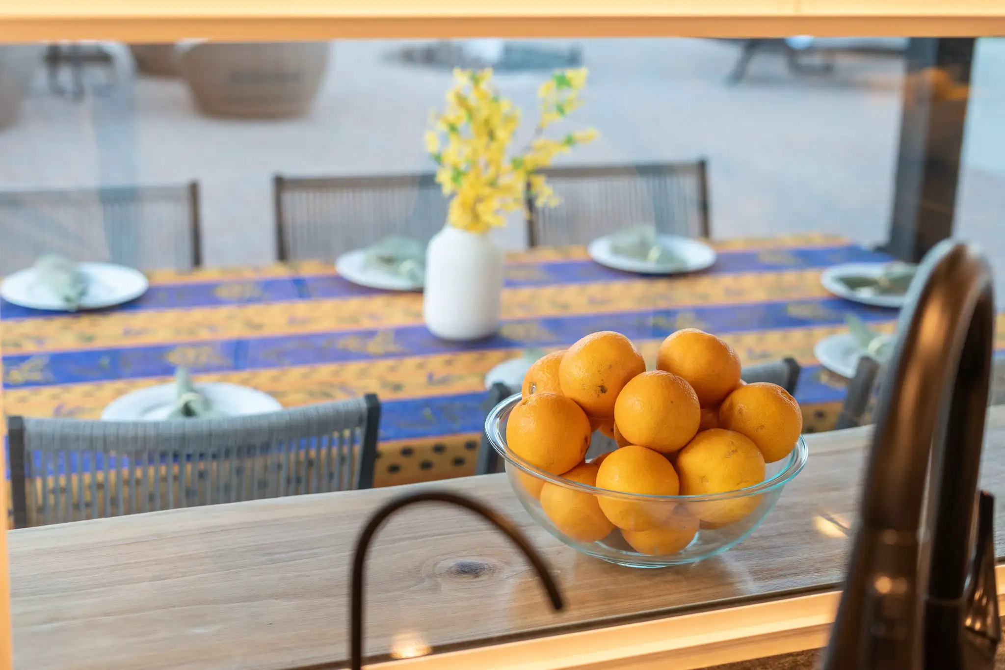 A bowl of oranges sits on a kitchen counter. In the background, an outdoor dining table is set with a bright blue and yellow cloth and a vase of yellow flowers.
