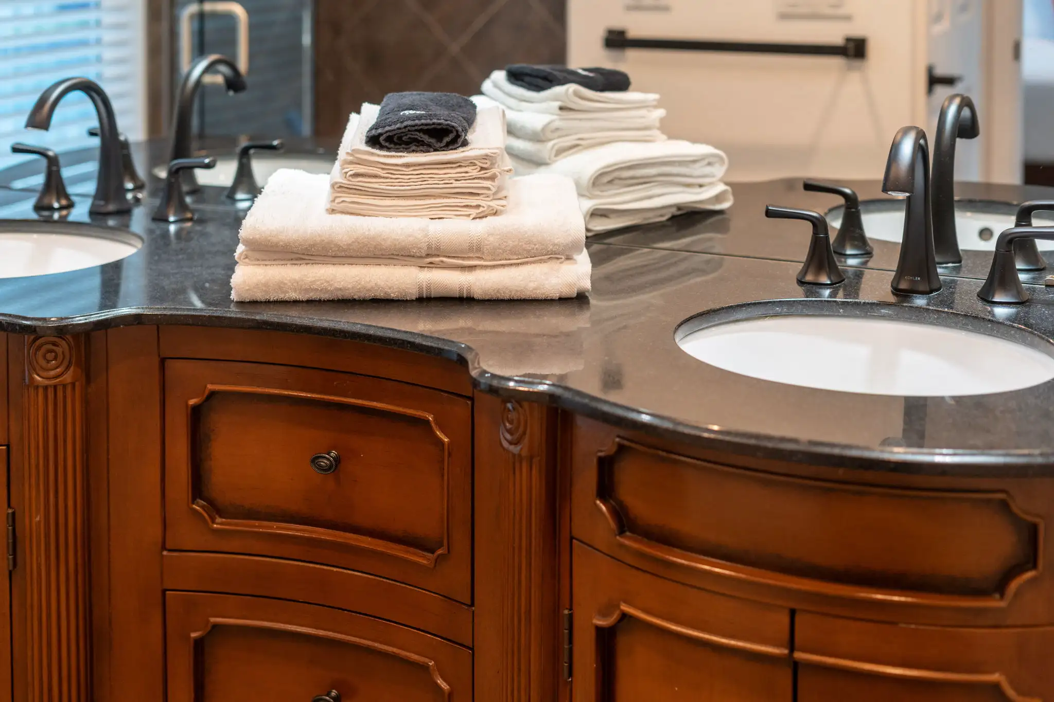 A luxurious double bathroom vanity featuring a dark wood cabinet, black granite counter, and modern bronze faucets. Clean, stacked towels sit between the two sinks.