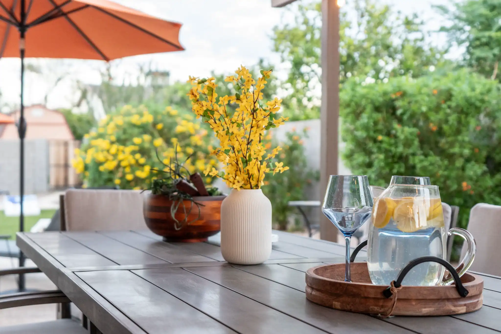 A gray slat-top patio table features a pitcher of lemon water and a vase of bright yellow flowers, set against lush green landscaping and an orange sun umbrella.