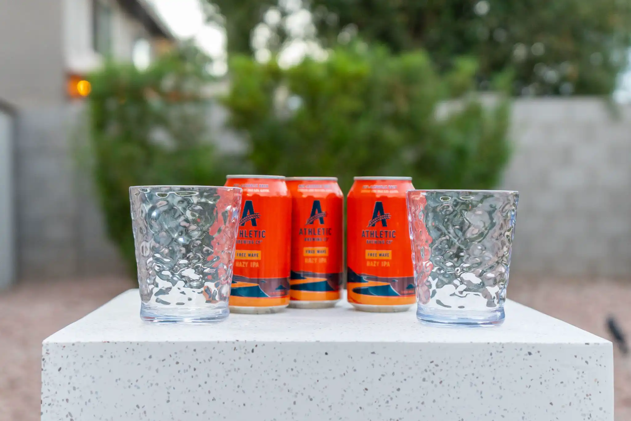 Three orange cans of Athletic Brewing Free Wave Hazy IPA and two textured glasses are displayed on a white outdoor counter.