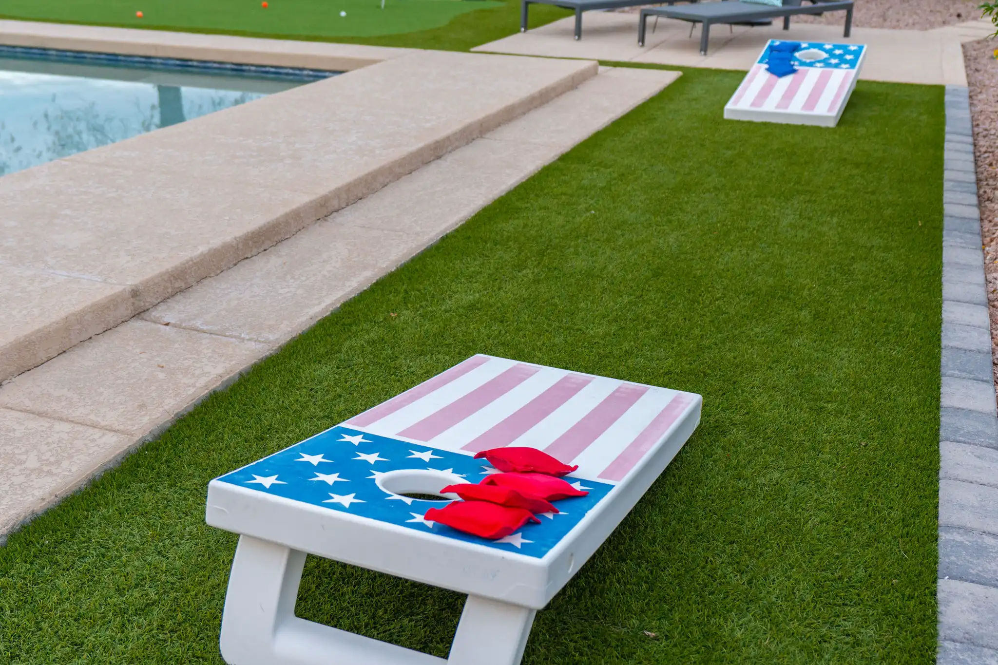 A cornhole game featuring American flag boards is set up on artificial green turf beside a swimming pool. Three red bean bags rest near the hole on the closest board.