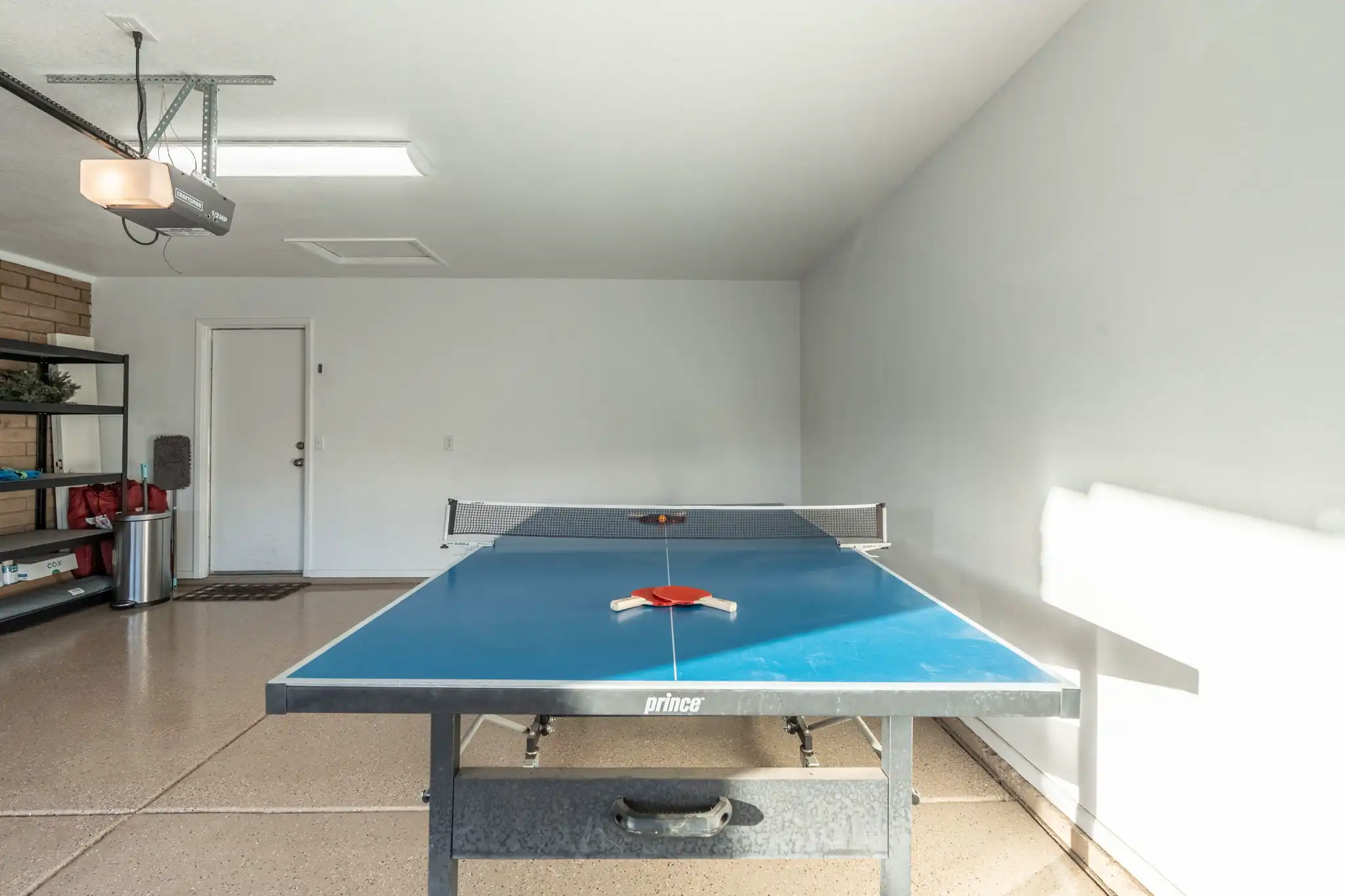 A blue ping pong table with two red paddles and a ball sits centered in a clean, brightly lit residential garage featuring white walls and a speckled floor.