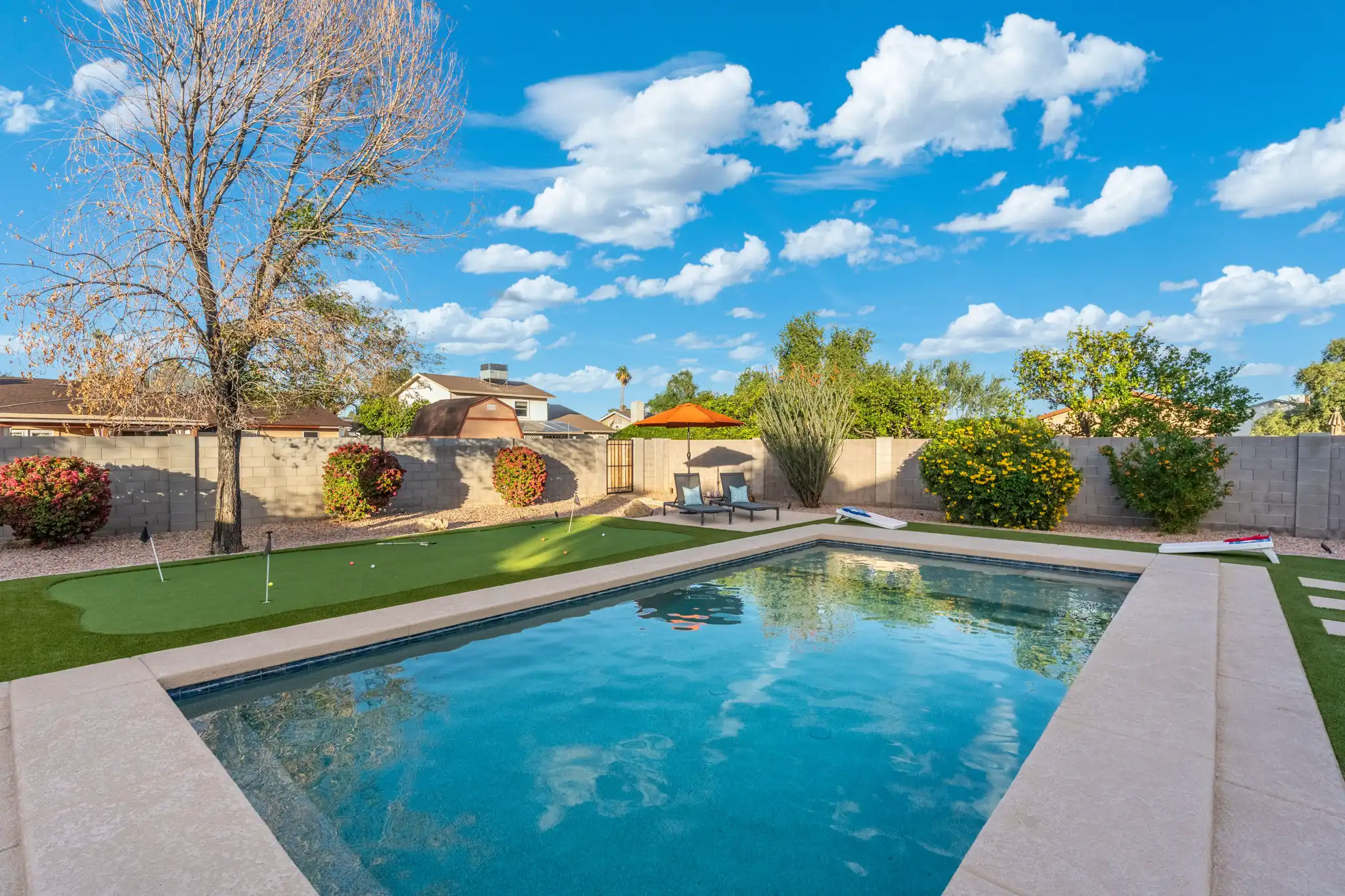 A bright backyard features a rectangular swimming pool next to a putting green with flags, dark lounge chairs, and cornhole boards, set against a sunny blue sky.