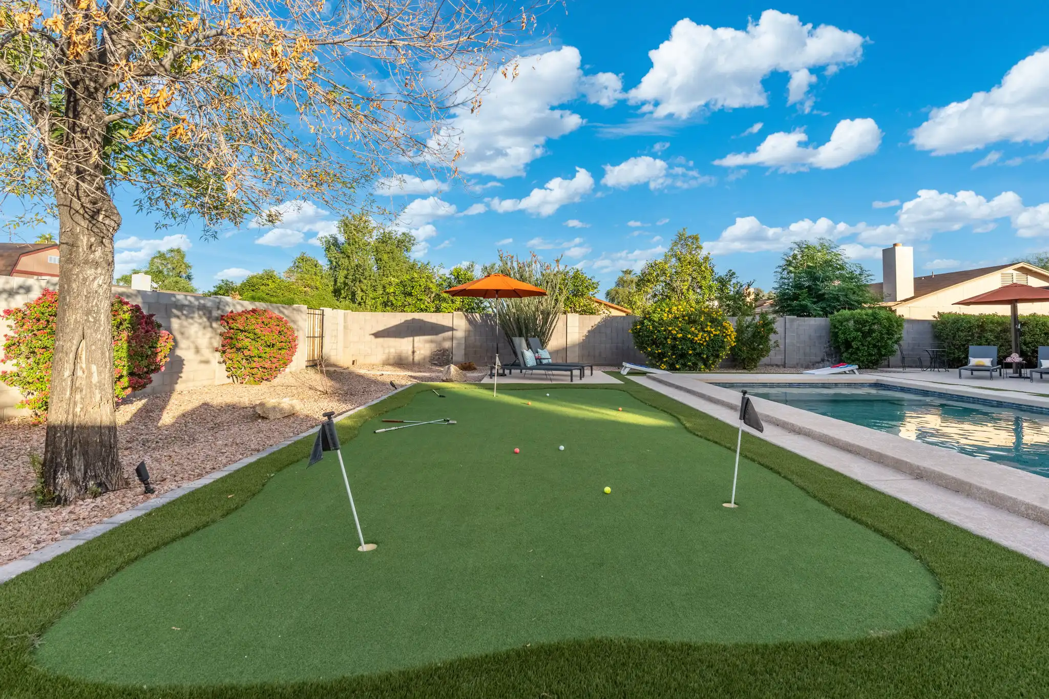 A sunny backyard shows a putting green with golf balls and flags next to a clear swimming pool. Lounge chairs and orange umbrellas are set up around the pool deck.