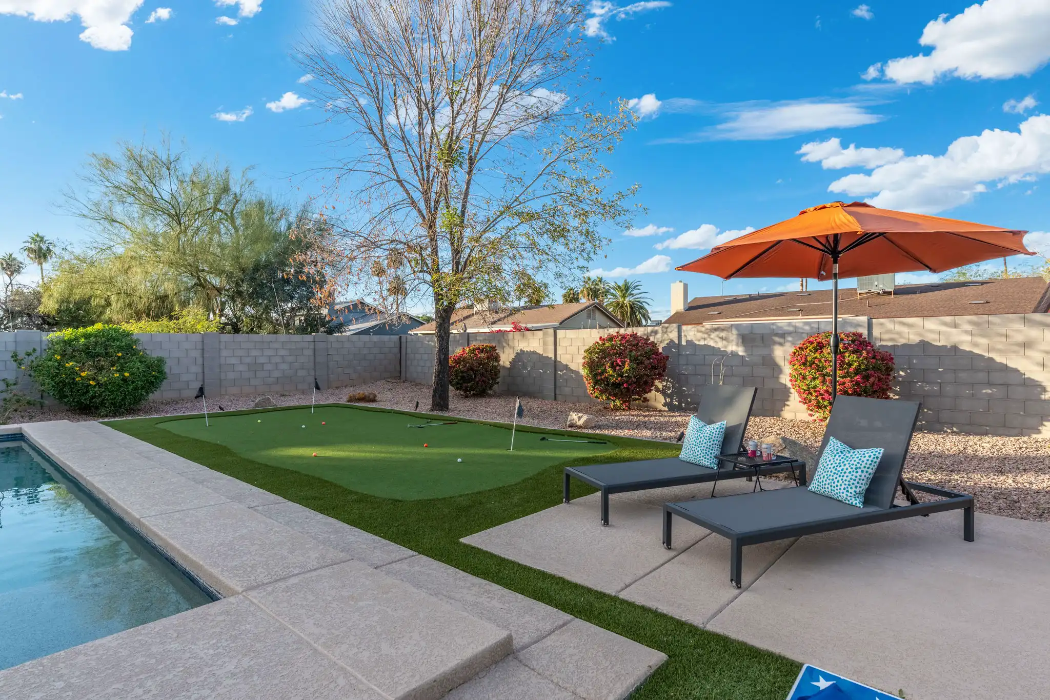 A vibrant backyard with a pool, a manicured putting green, and two grey lounge chairs shaded by a large orange umbrella.