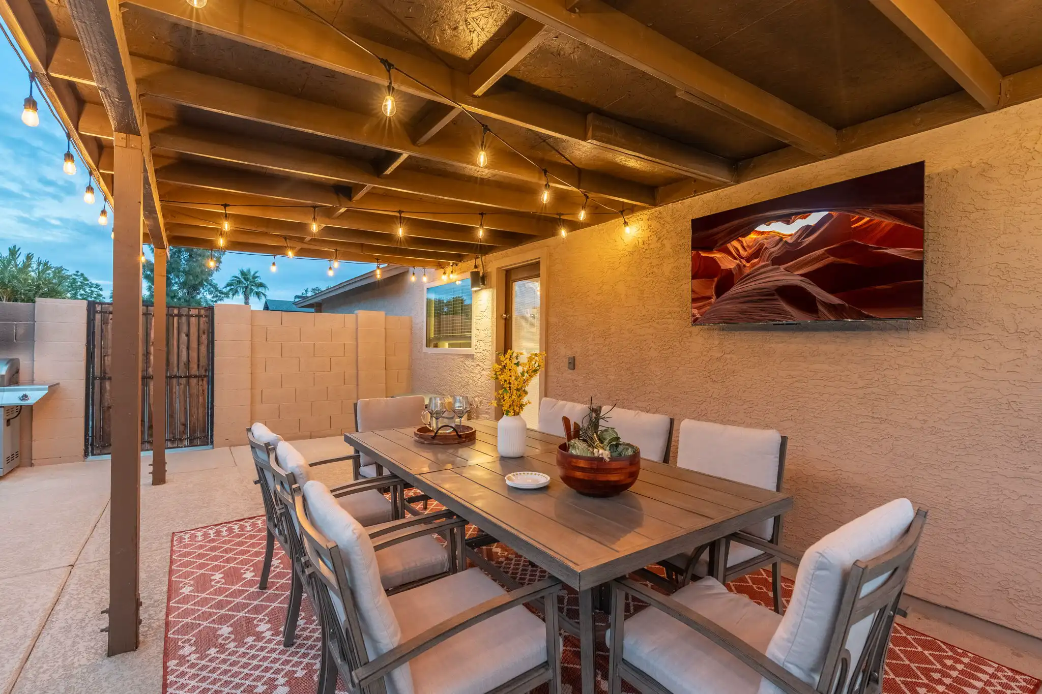 A covered outdoor patio illuminated by string lights features a wooden dining table and cushioned seating for eight, with a TV mounted on the wall.
