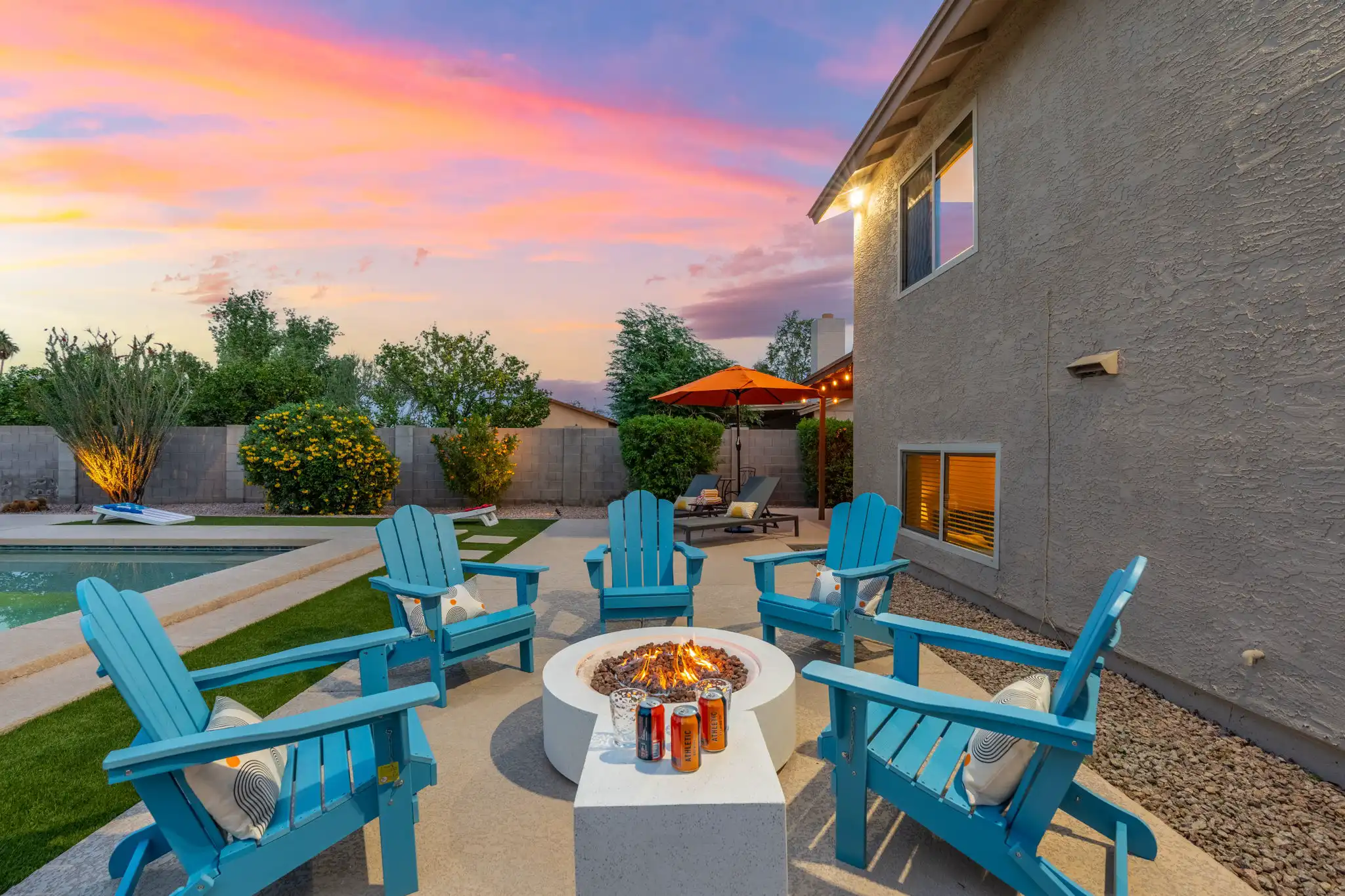 A residential backyard patio at sunset featuring a lit fire pit surrounded by five bright blue Adirondack chairs. A swimming pool and an orange umbrella are visible under a vibrant sky.