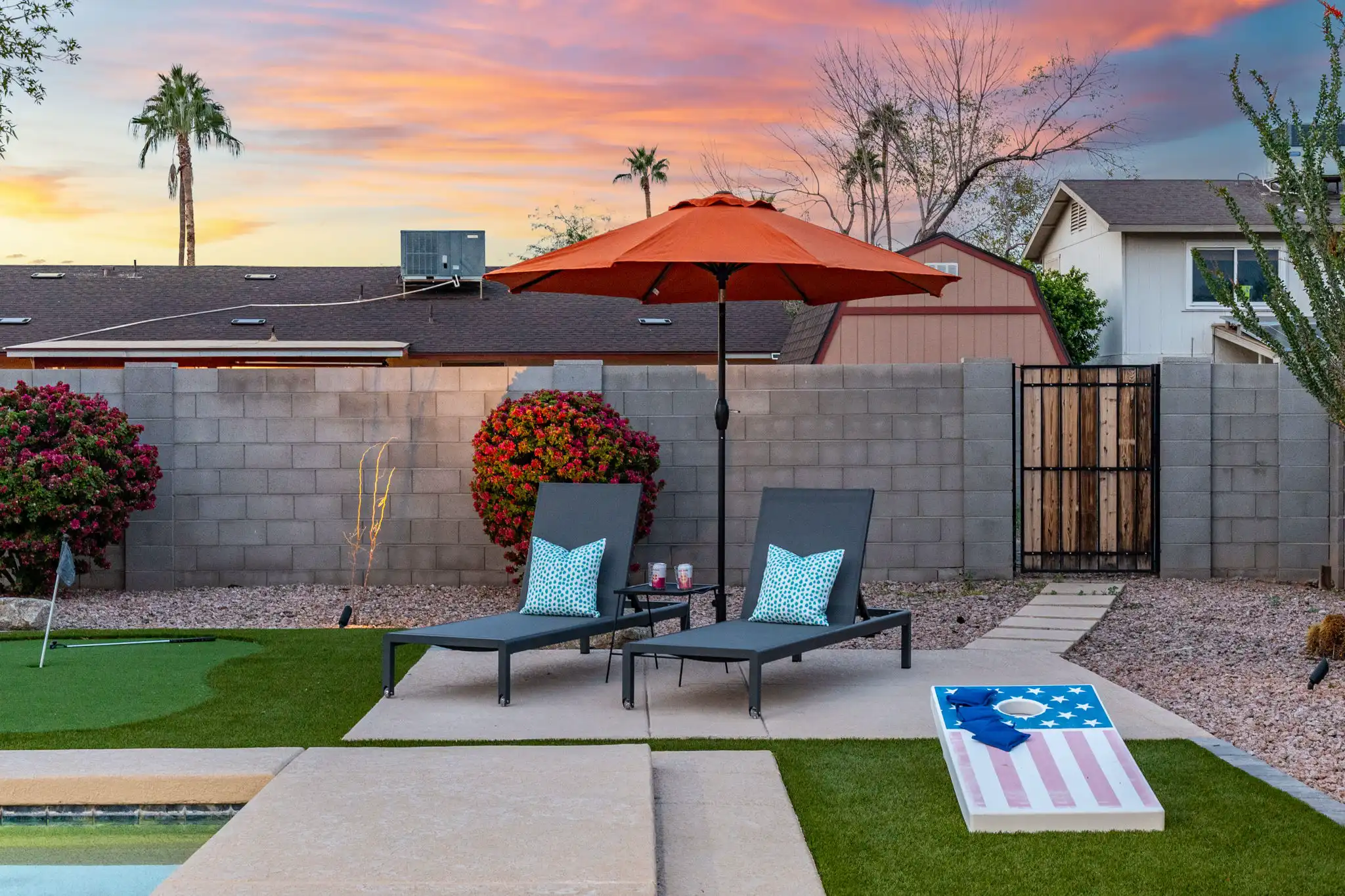 Two grey chaise lounges with bright pillows sit under an orange umbrella next to a cornhole game. The backyard features artificial turf, a pool edge, and a dramatic sunset.