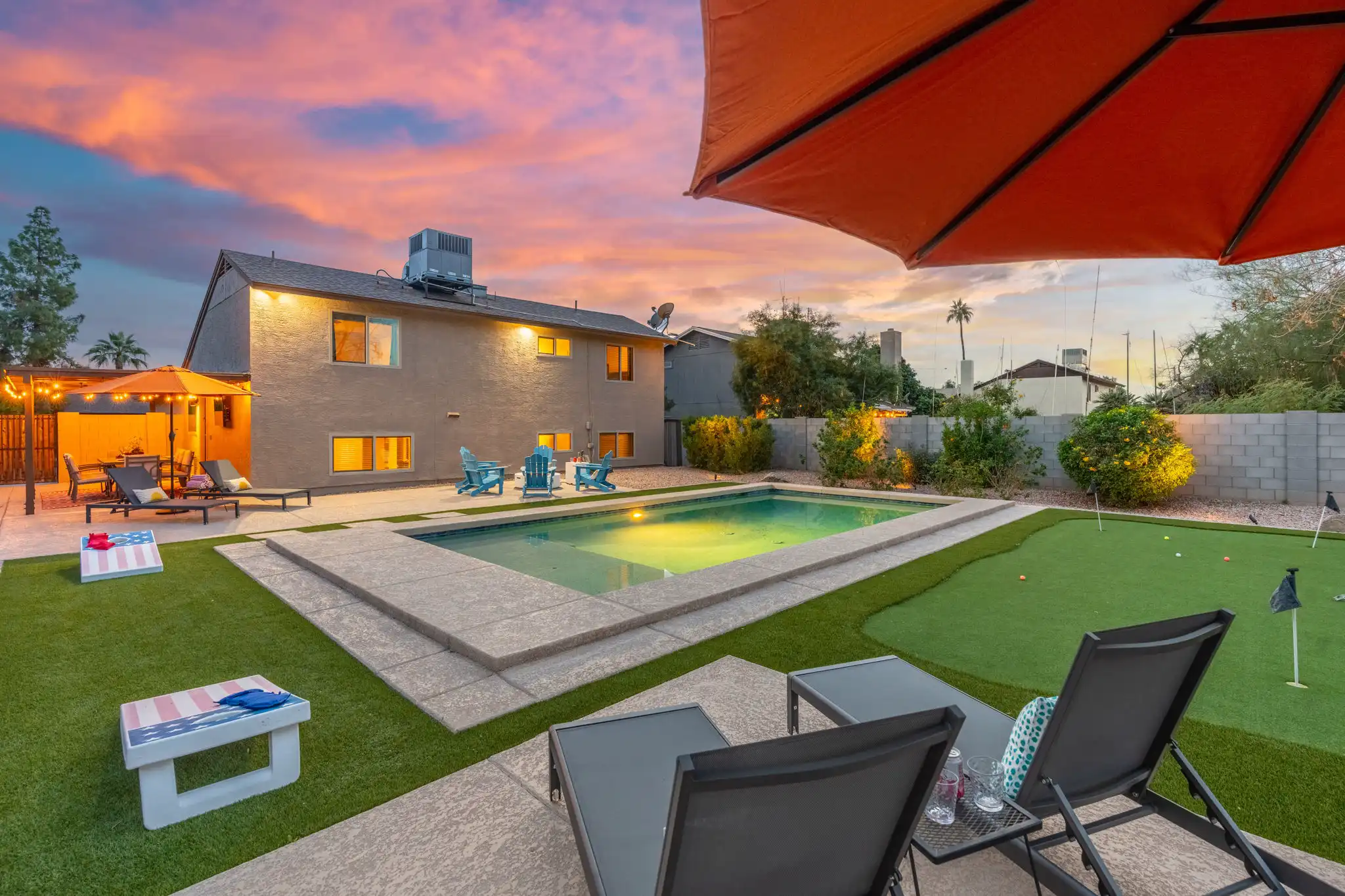 A colorful sunset illuminates a private backyard with a swimming pool, lounge chairs, a putting green, and outdoor games.