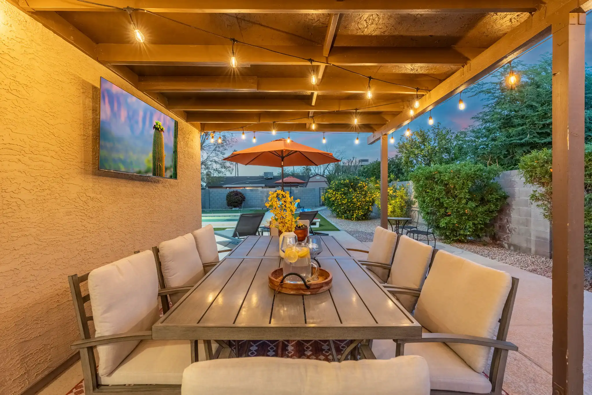 A covered patio dining area with string lights and a mounted TV. The long table is set for dinner with drinks, surrounded by comfortable cushioned chairs.