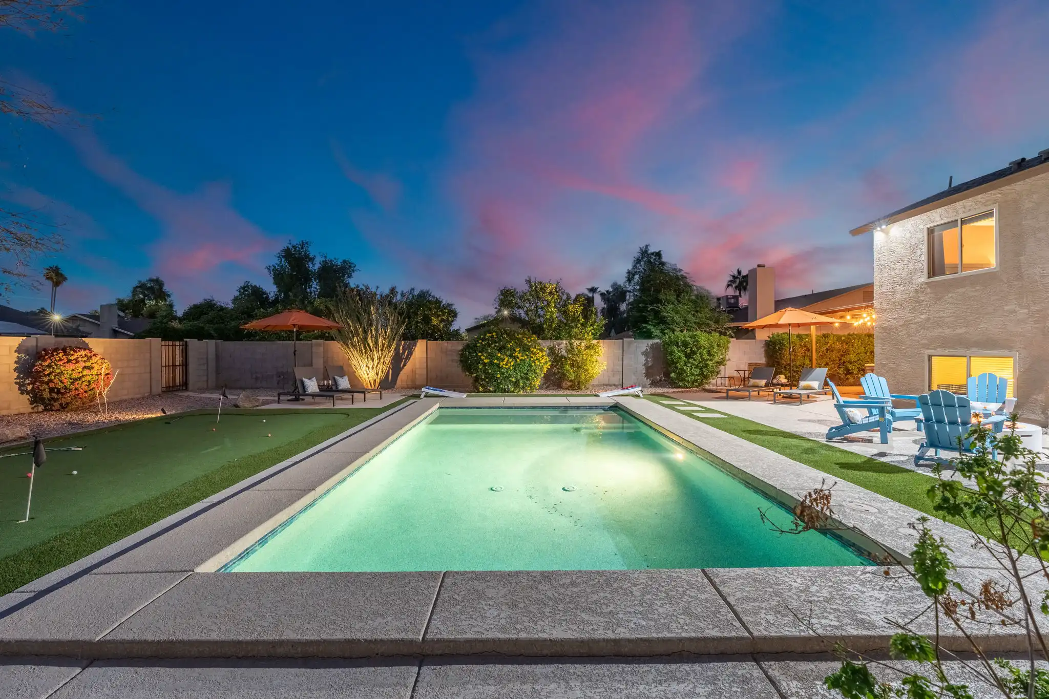 An exterior view of a luxury residential backyard at dusk, featuring a lit pool, a putting green, and a patio with blue chairs and string lights under a dramatic blue and pink sky.