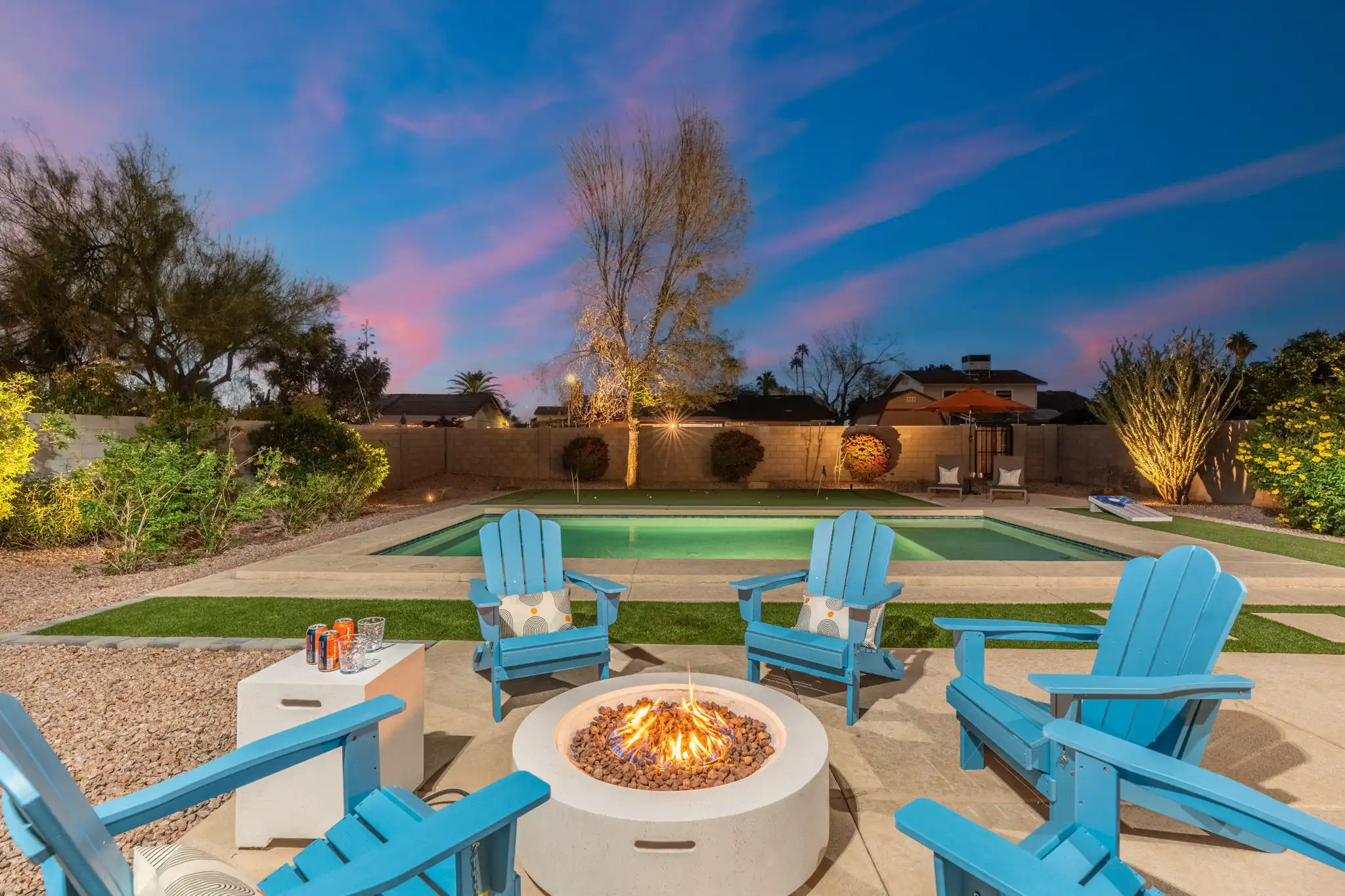 A vibrant backyard scene at twilight with a lit fire pit and five blue Adirondack chairs, situated on a patio overlooking a swimming pool and a small putting green.