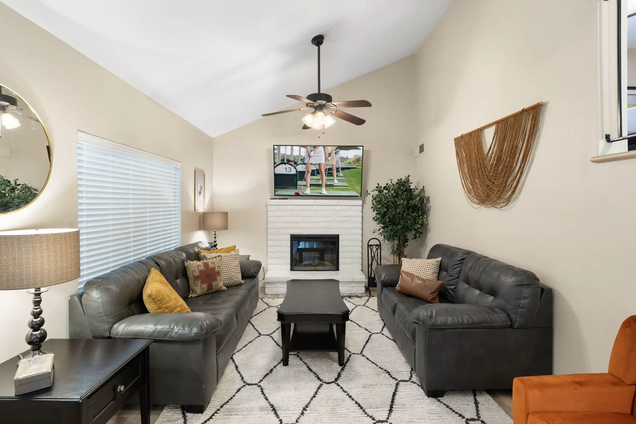 A bright living room features a vaulted ceiling, two dark leather sofas, and a black coffee table on a geometric rug. A TV is mounted above the white brick fireplace.