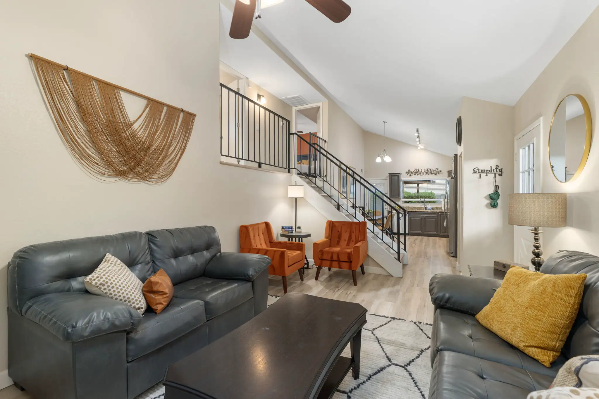 A bright, open-concept living space with dark leather sofas, orange armchairs, and a central staircase leading to a loft. A kitchen is visible in the background.