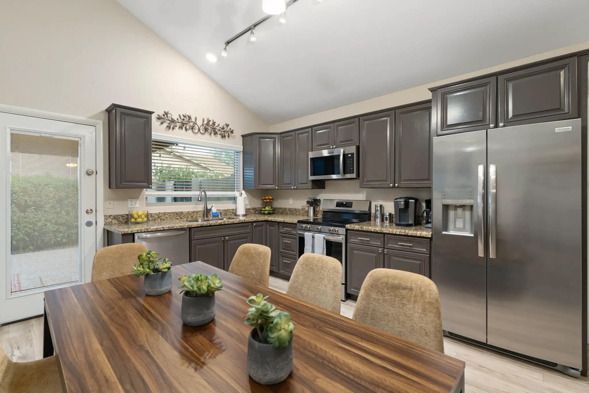 A modern kitchen and dining area with vaulted ceilings. The kitchen has dark gray cabinets and stainless steel appliances; a wooden table with potted plants is in the foreground.