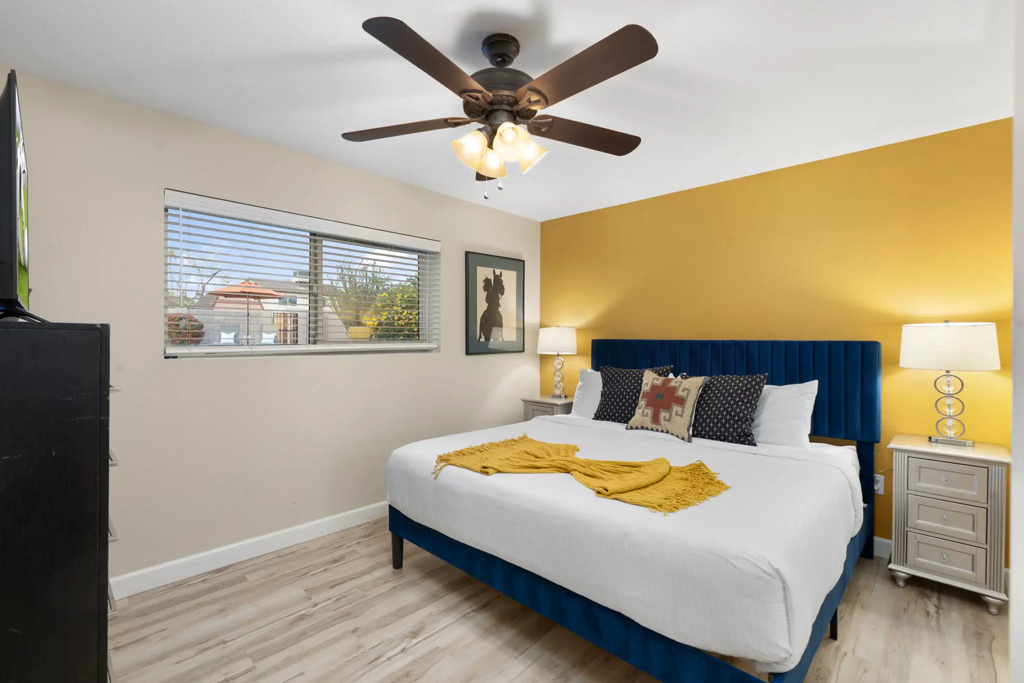 A modern bedroom featuring a king-sized bed with a blue velvet headboard, white linens, and a bright yellow accent wall. Silver nightstands flank the bed.