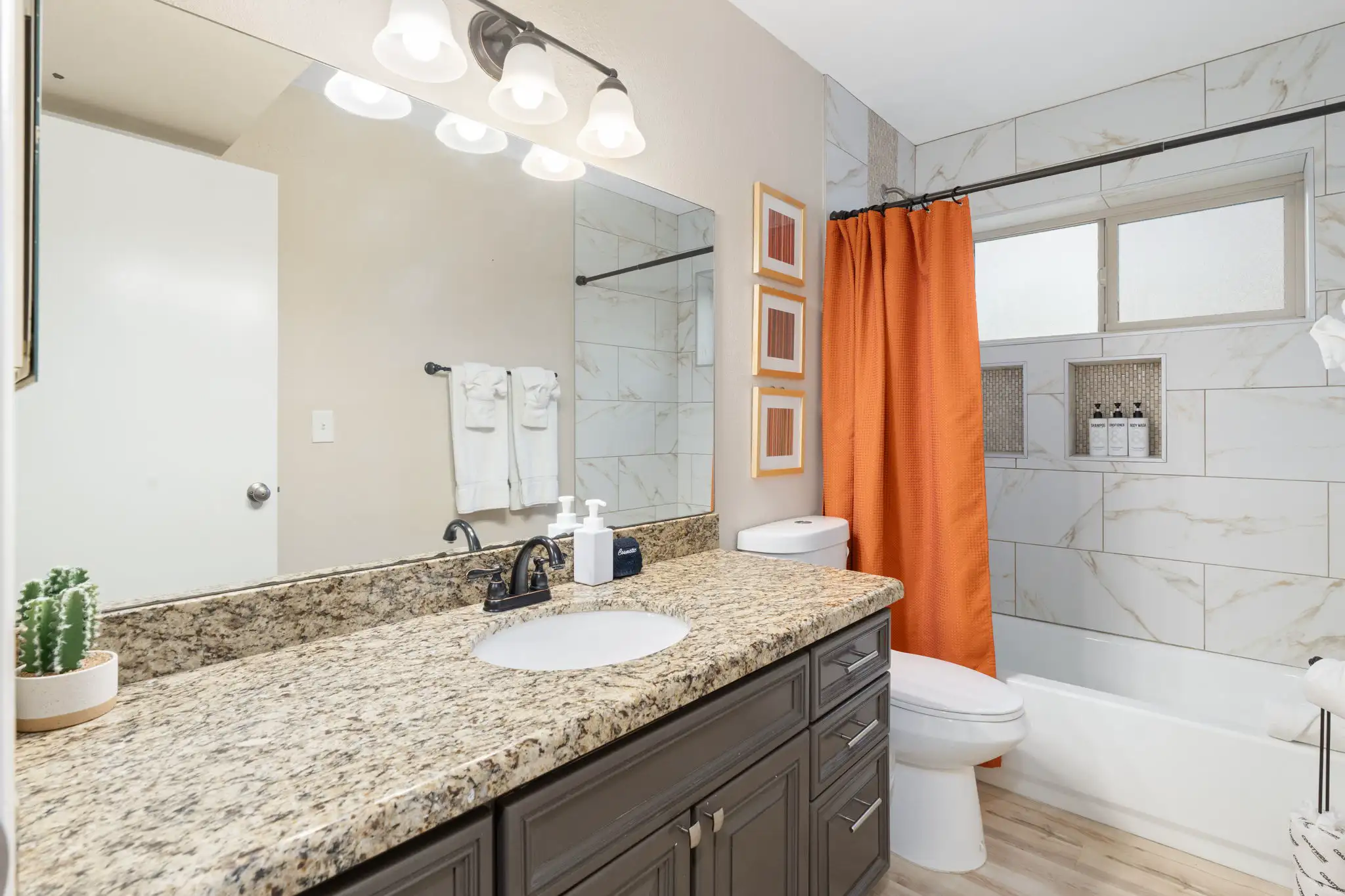 A modern bathroom features a dark vanity with a granite countertop and a white tub/shower area tiled in marble-look ceramic, accented by a bright orange shower curtain.