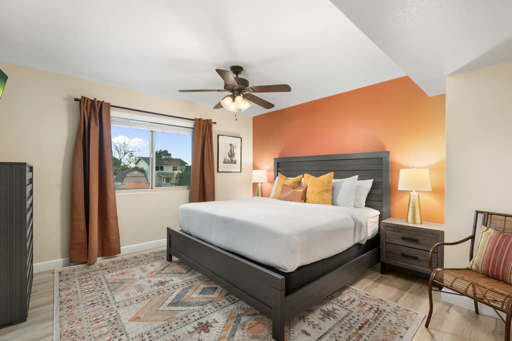 A modern bedroom with a dark gray bed, a warm copper accent wall, and a patterned area rug. A ceiling fan and a window overlooking houses complete the space.