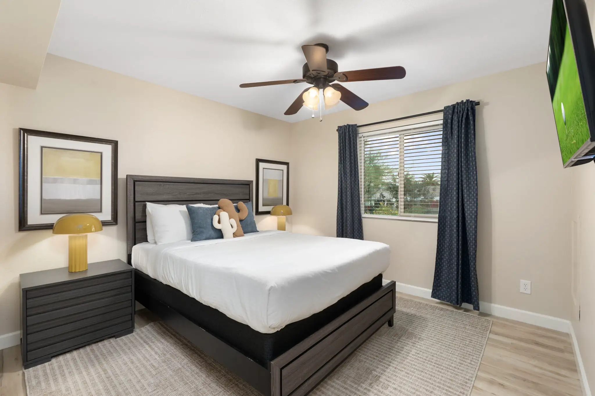 A modern bedroom features a dark gray wood bed frame, nightstand, and matching ceiling fan. The bed has a white comforter and a cactus pillow. Yellow lamps sit on the nightstands next to abstract art.