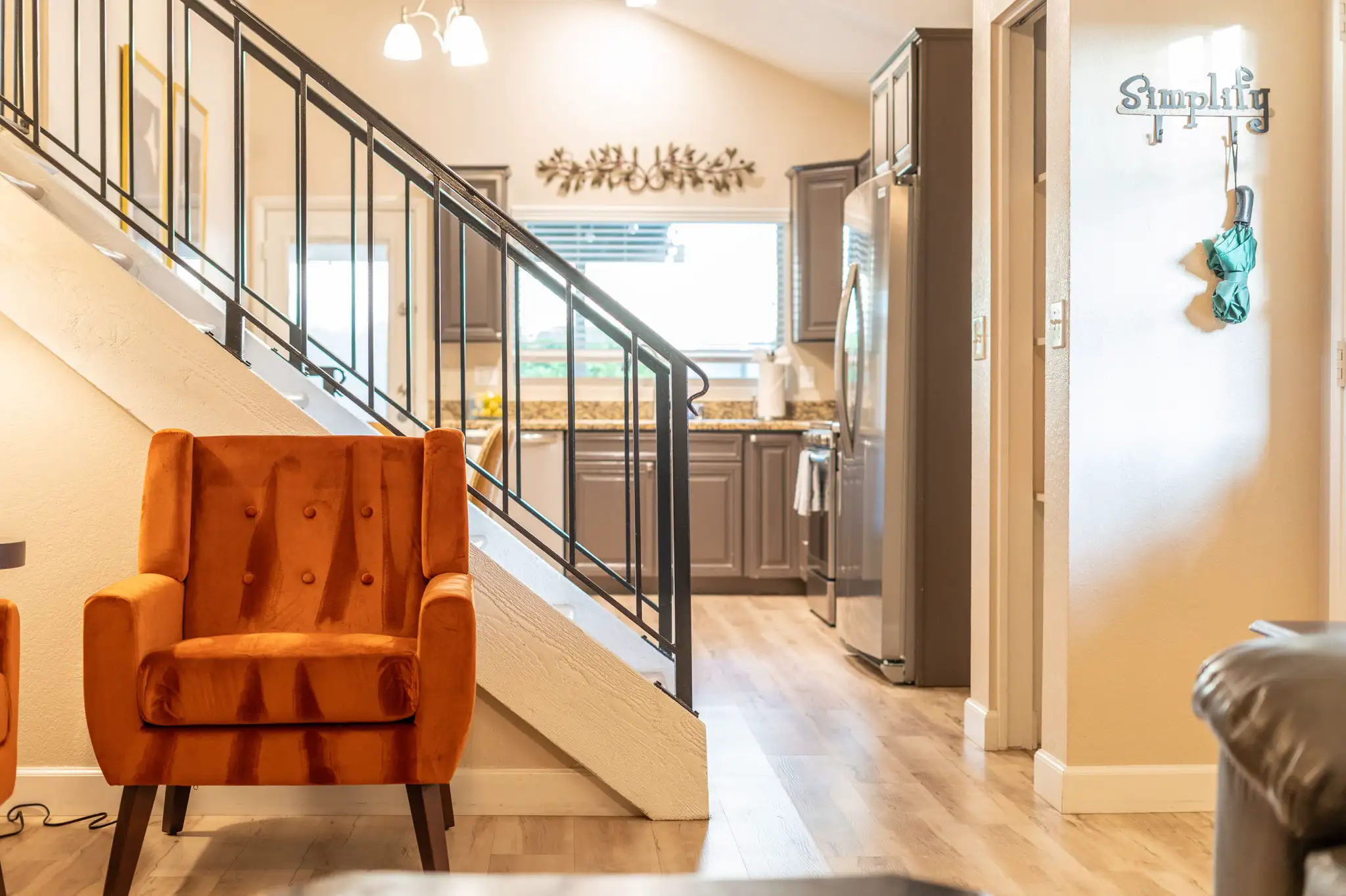 An orange velvet armchair sits near a staircase with iron railings, opening to a kitchen featuring gray cabinets and stainless steel appliances.