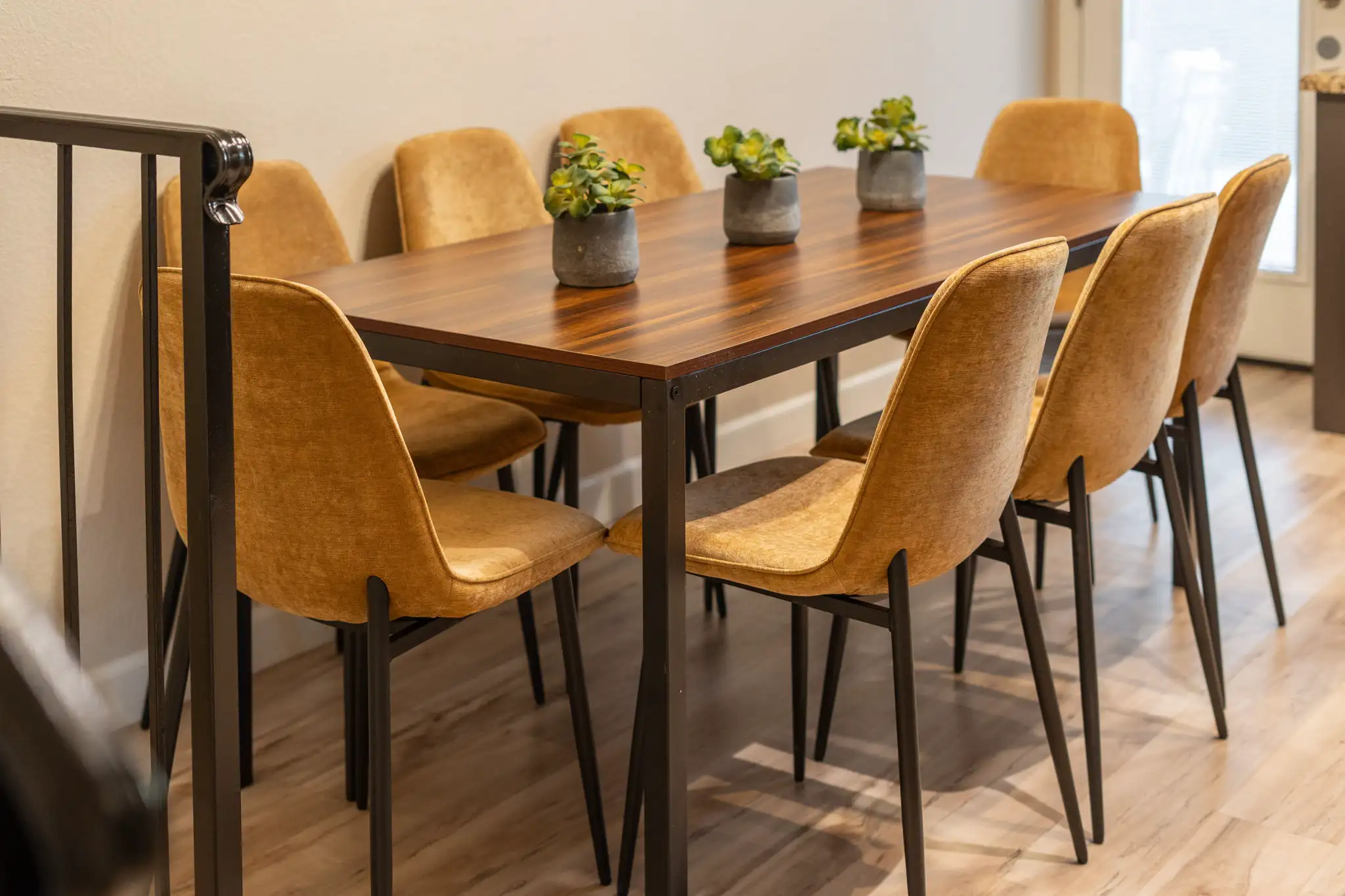 A modern dining table with a dark wood top and black legs. The table is surrounded by mustard yellow chairs and holds three small potted plants.
