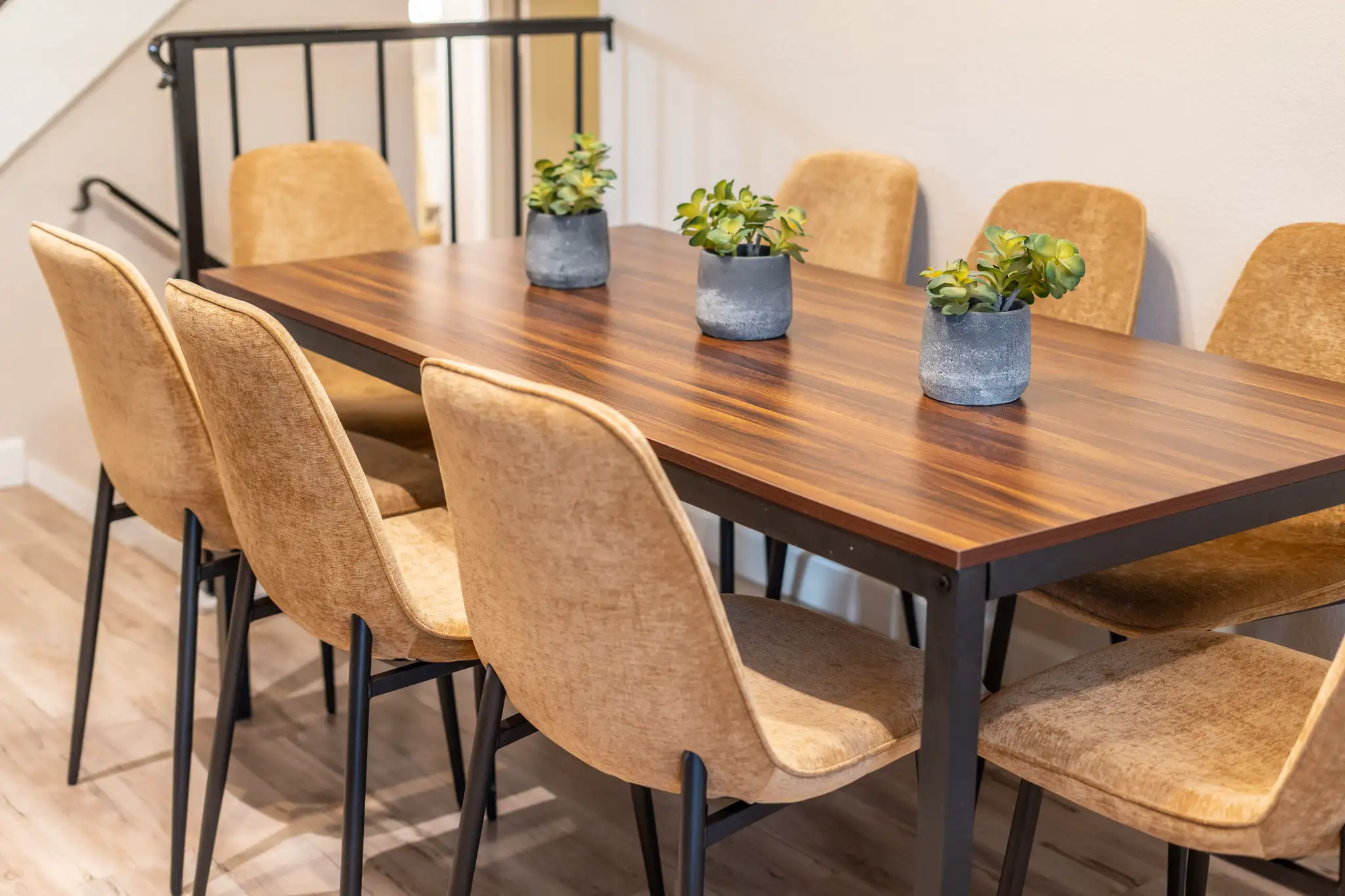A modern dining table with a dark wood top and six plush tan chairs in a home setting. Three small potted plants sit on the table.