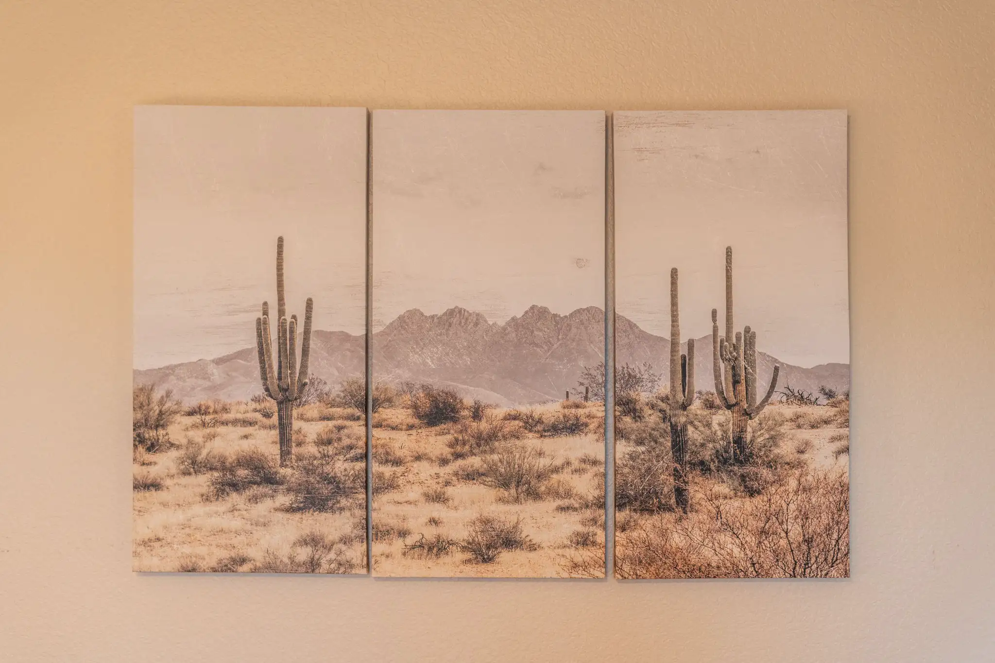 A triptych canvas artwork displays a muted desert scene with prominent saguaro cacti in the foreground, scrub brush, and distant mountains.