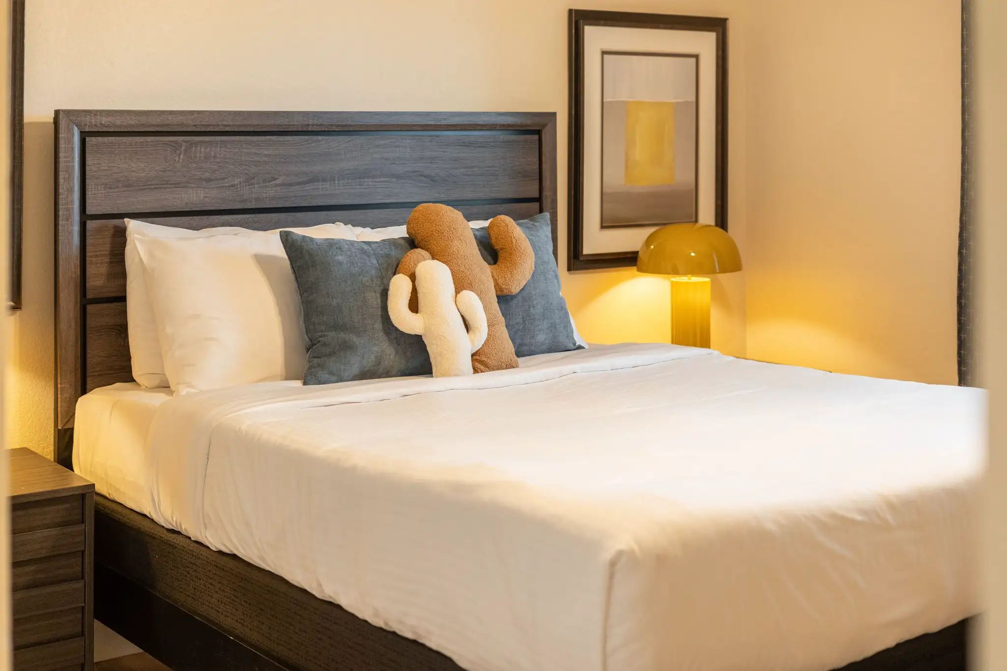 A cozy bedroom interior with a dark wooden bed frame, white bedding, and two decorative cactus-shaped pillows next to a warm yellow lamp.