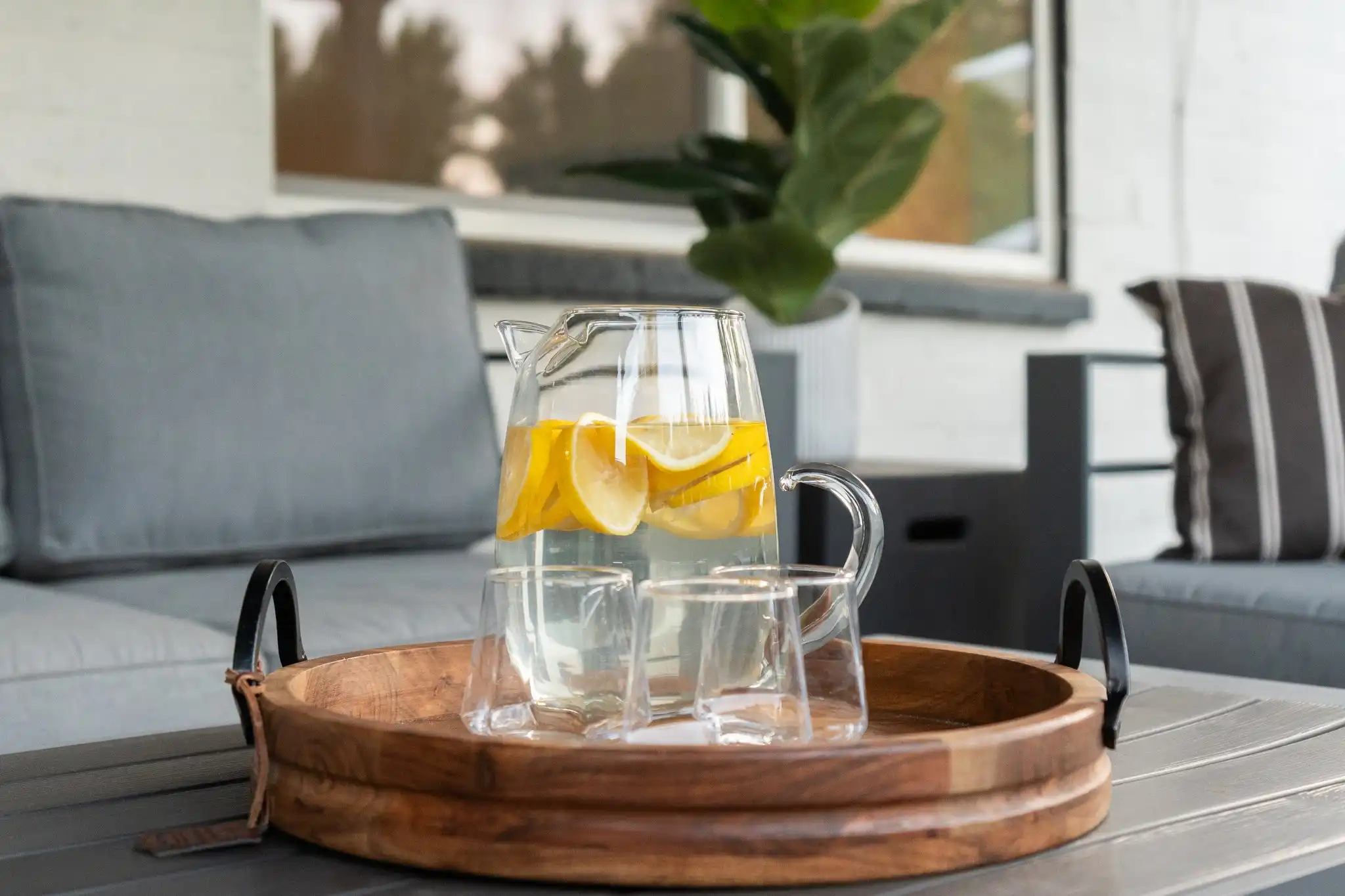 A pitcher of lemon-infused water and two drinking glasses rest on a round wooden tray on a dark outdoor table. Gray patio furniture is visible behind it.